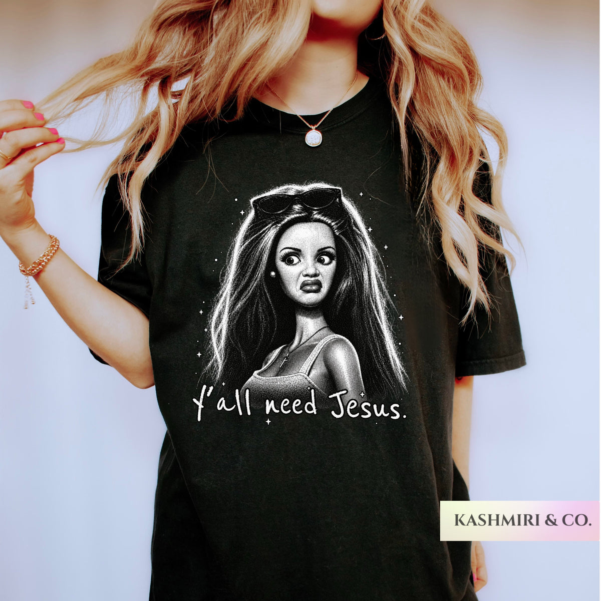 A woman wearing a black graphic t-shirt with a black and white illustration of a woman's face and the text 'Y'all need Jesus.' The brand name 'KASHMIRI & CO.