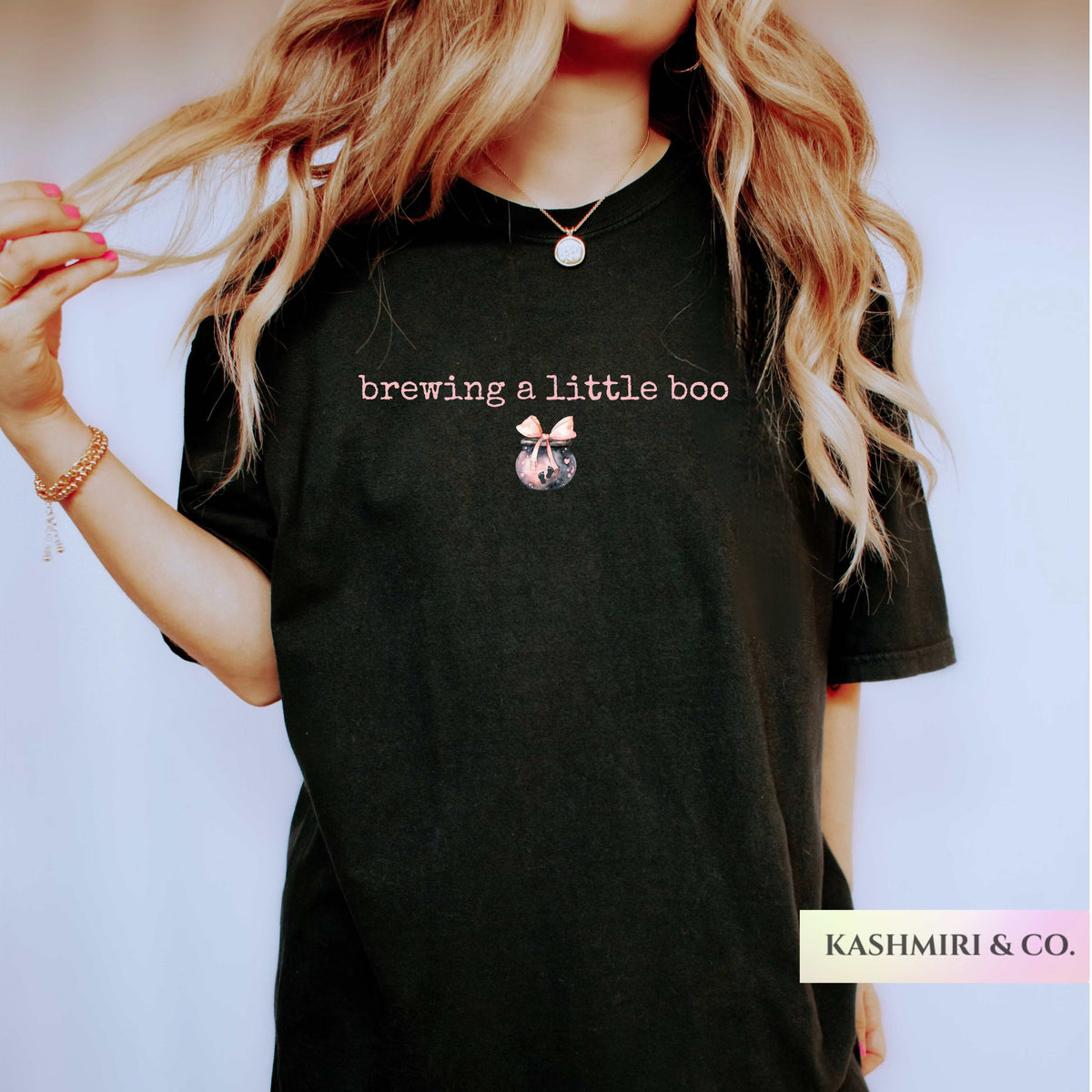A woman wearing a black graphic t-shirt with the text 'brewing a little boo' and a small potion brew illustration. The brand name 'KASHMIRI & CO.