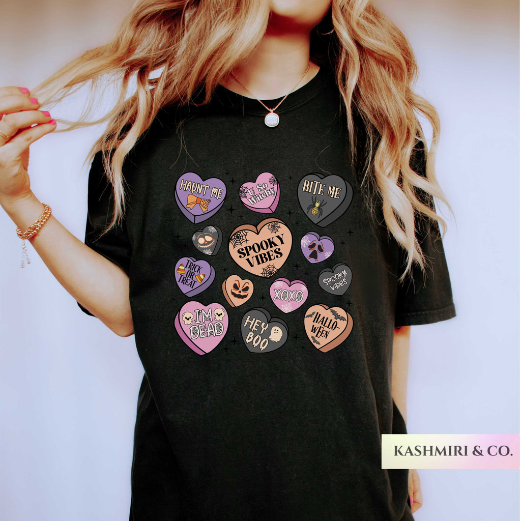 A woman wearing a black graphic t-shirt with multiple colorful heart-shaped patches featuring Halloween-themed phrases like 'TRICK or TRAITOR', 'SPOOKY VIBES', and 'HE' BOO'.