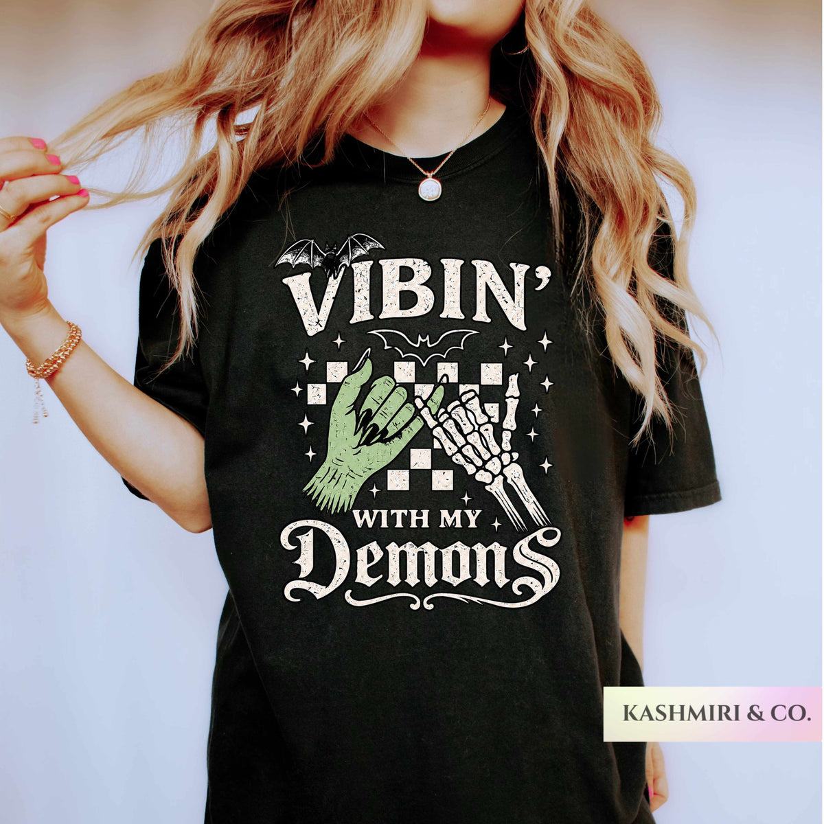 A woman wearing a black graphic t-shirt with a gothic design that reads 'VIBIN' WITH MY DEMONS' featuring a green zombie hand and a skeleton hand. The brand name 'KASHMIRI & CO.