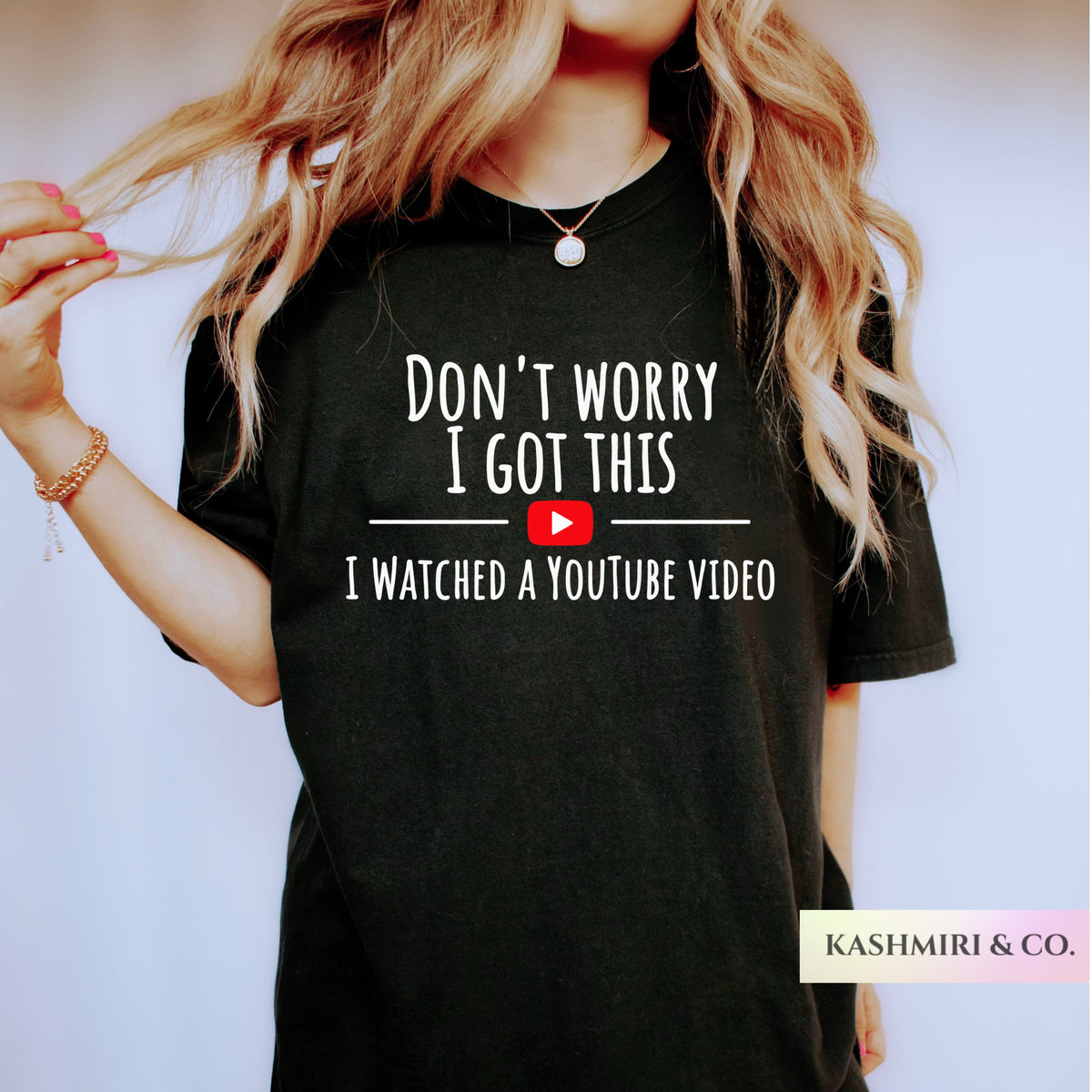 A woman wearing a black graphic t-shirt with the text 'DON'T WORRY I GOT THIS I WATCHED A YOUTUBE VIDEO' printed in white, featuring a red YouTube play button icon. The brand name 'KASHMIRI & CO.