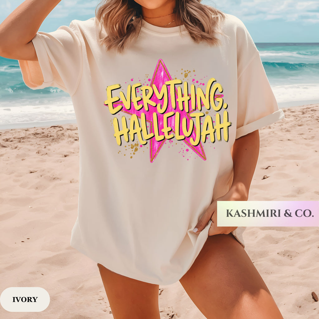 A woman wearing an oversized ivory graphic t-shirt with a pink star and yellow text reading 'EVERYTHING, HALLELUJAH' on a sandy beach with the ocean in the background. A brand label 'KASHMIRI & CO.