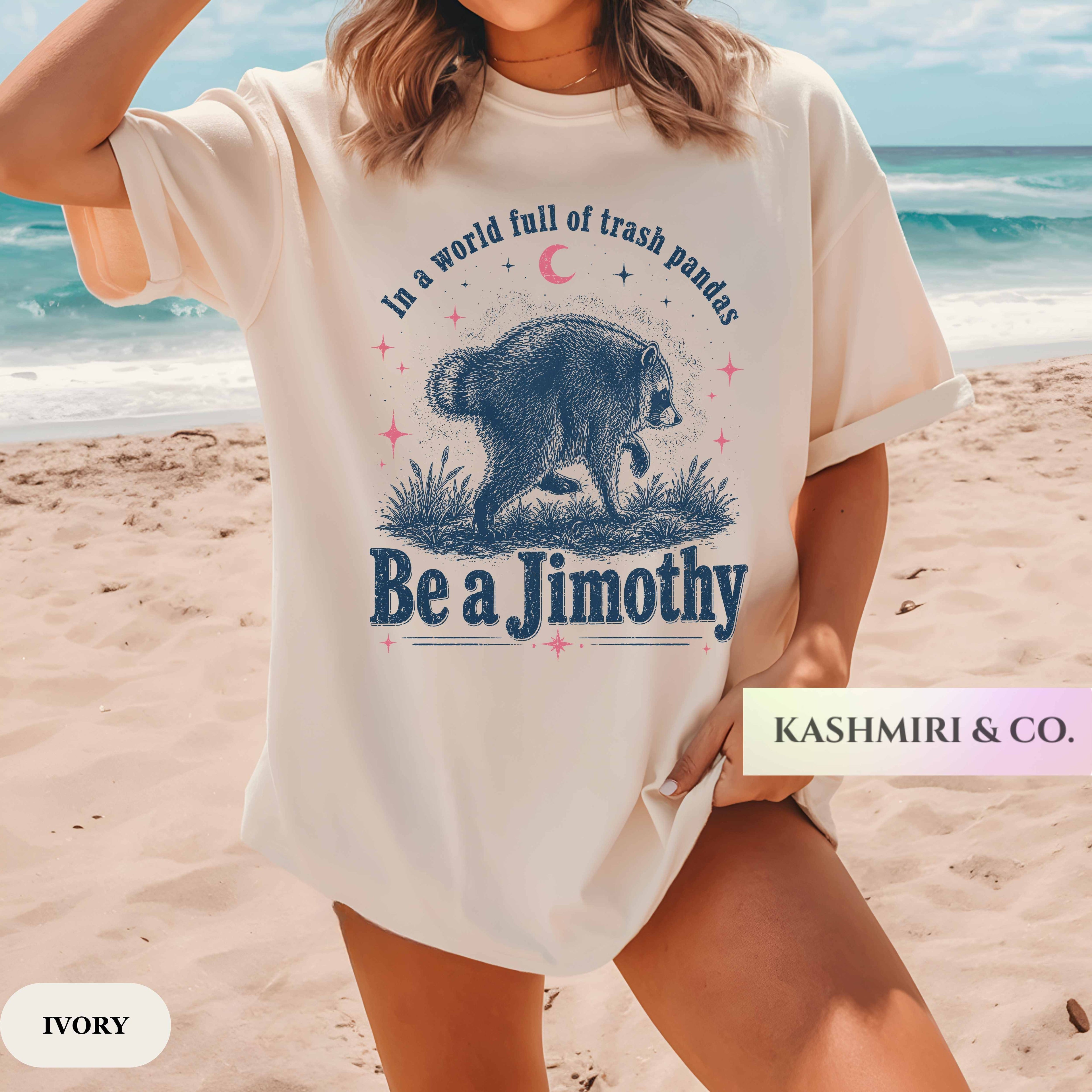 A woman on a sandy beach wearing an oversized ivory graphic T-shirt with a blue bear illustration and the text 'In a world full of trash pandas Be a Jimothy'. A 'KASHMIRI & CO.