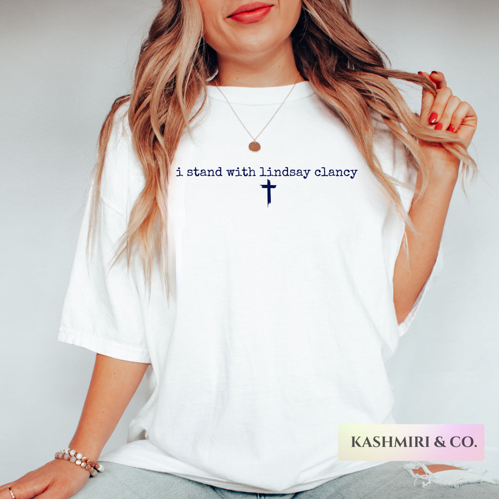 A woman wearing a white oversized T-shirt with the printed message 'i stand with lindsay clancy' and a small cross in navy blue on the chest. The photo is cropped from the mouth down, showing her hair, necklace, bracelet, and jeans against a light gray studio background.