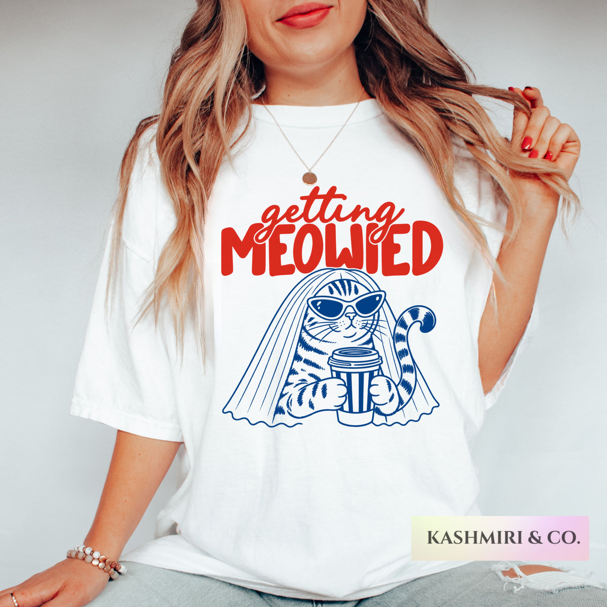 A woman wearing a white graphic t-shirt with a blue and red print that reads 'getting MEOWIED' above an illustration of a cat in a bridal veil holding a coffee cup. The brand name 'KASHMIRI & CO.