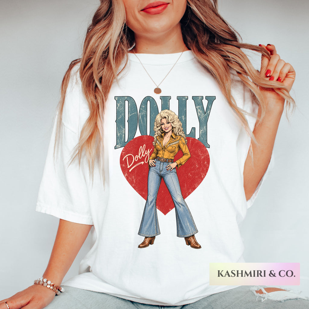 A woman wearing a white graphic t-shirt featuring a vintage-style illustration of Dolly Parton with the text 'DOLLY' and a red heart. The photo is cropped from the chin down, set against a plain light gray background.