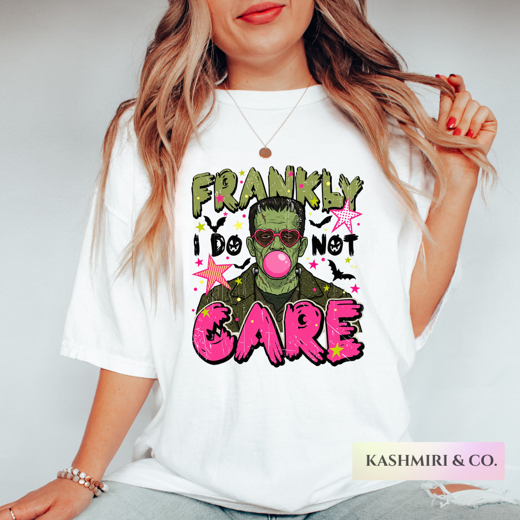 A woman wearing a white graphic t-shirt with a Frankenstein-themed print that reads 'FRANKLY I DO NOT CARE' in green, pink, and black lettering. The brand name 'KASHMIRI & CO.