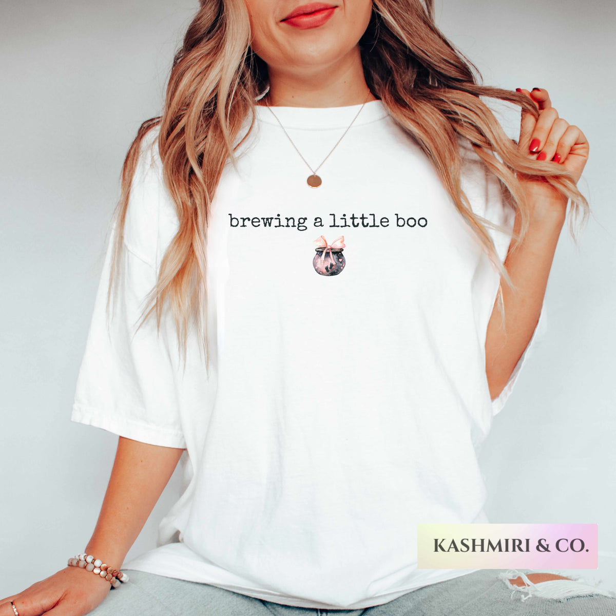 A woman wearing a white oversized graphic t-shirt with the text 'brewing a little boo' and a small potion brew illustration with a 'KASHMIRI & CO.