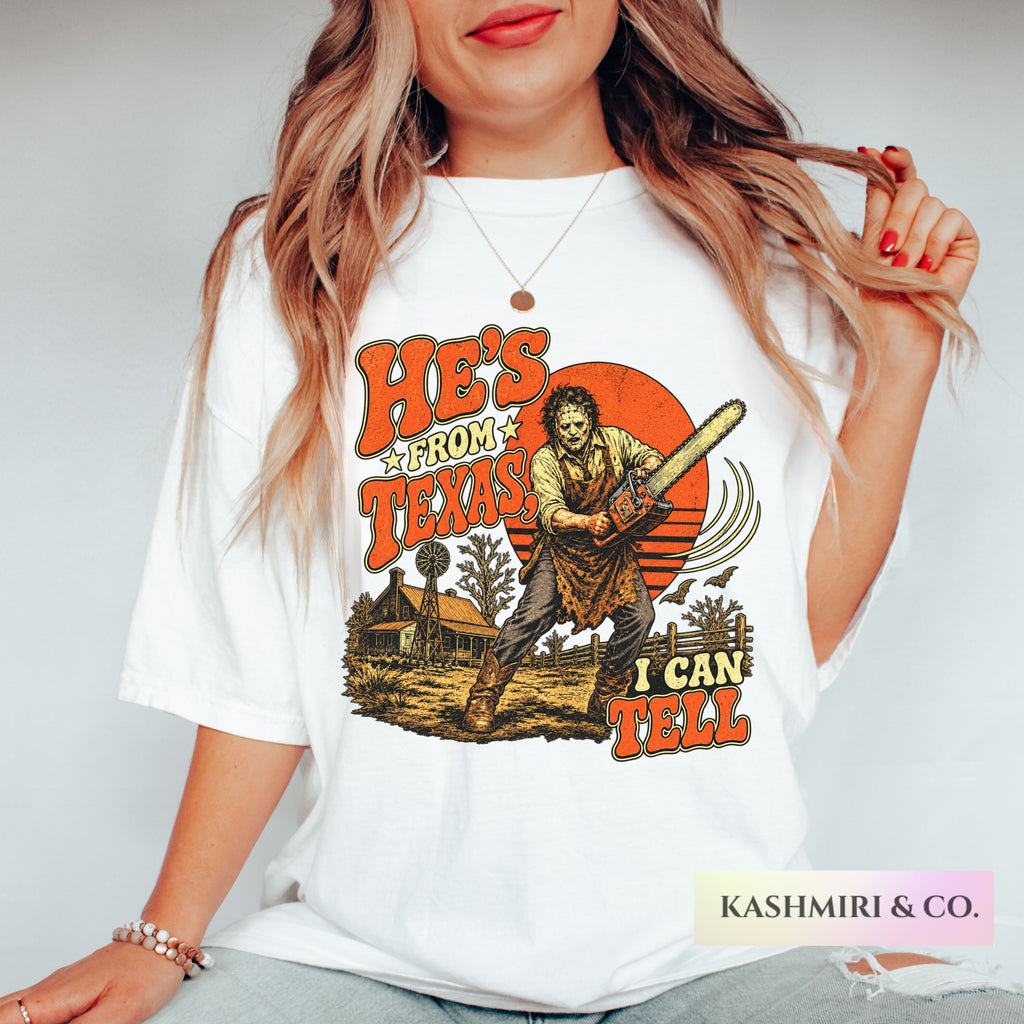 A woman wearing a white graphic t-shirt with a horror-themed print featuring Leatherface and the text 'HE'S FROM TEXAS I CAN'T TELL'. The photo is cropped from the chin down, showing the fit of the shirt.