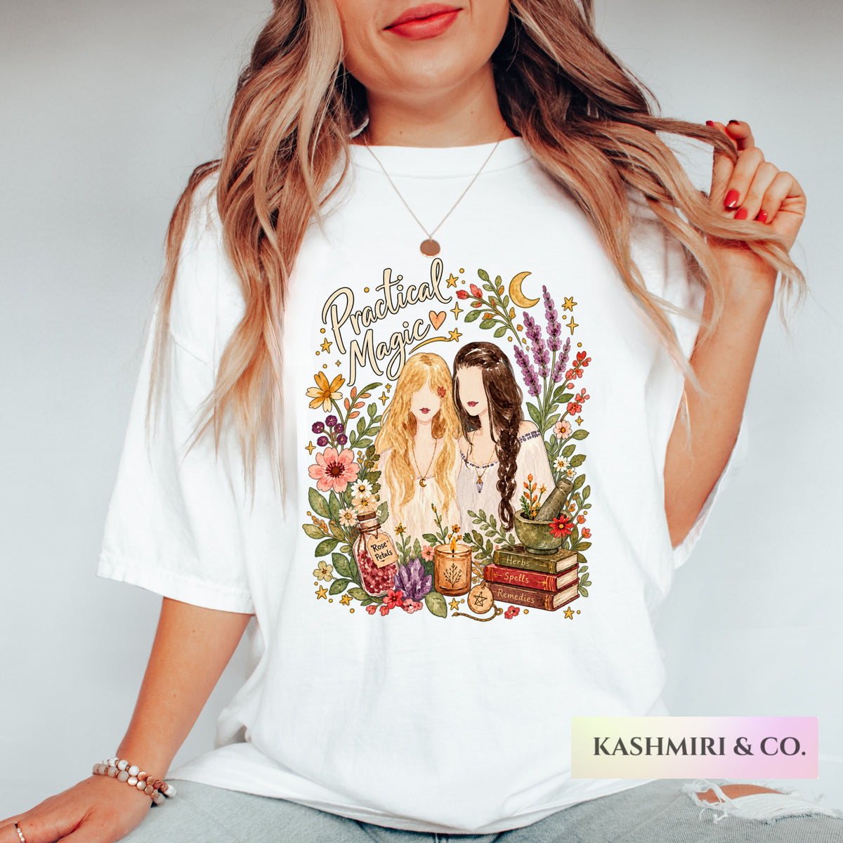 A woman wearing a white graphic t-shirt with a colorful illustration of two girls surrounded by flowers, books, and the words 'Practical Magic'.