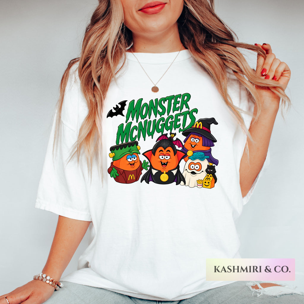 A woman wearing a white graphic t-shirt with a colorful 'Monster McNuggeds' cartoon design, posed against a light gray background.