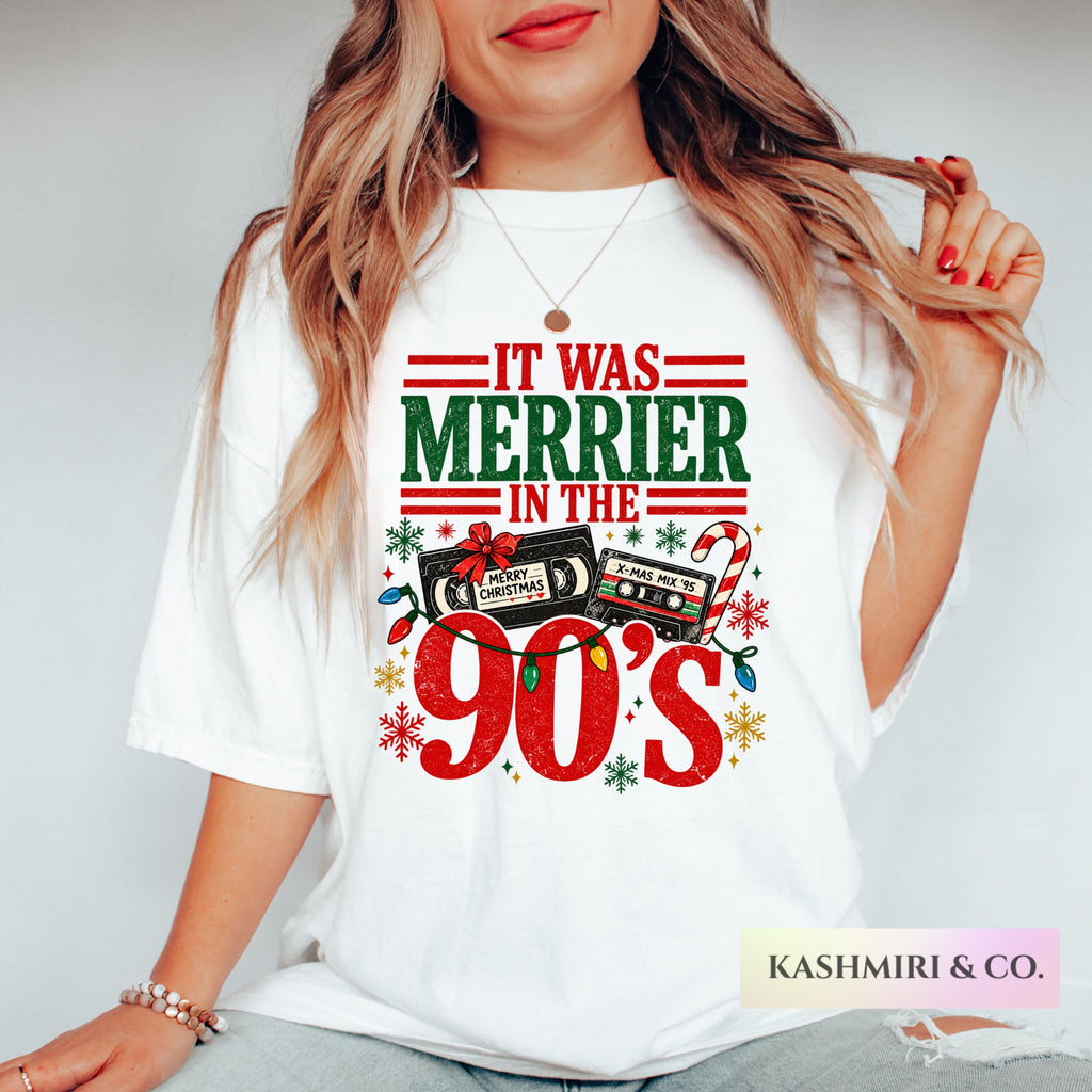 A woman wearing a white graphic t-shirt with a festive 90s Christmas design that reads 'IT WAS MERRIER IN THE 90's' with cassette tapes, candy canes, and snowflakes. The photo is cropped from the chin down, set against a plain light gray background.