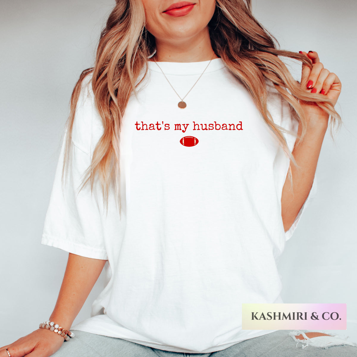 A woman wearing a white oversized t-shirt with red text that reads 'that's my husband' taylor swift travis kelce and a small red football graphic on the chest. The photo is cropped from the mouth down, showing her hair, hand, jewelry, and jeans against a light gray background.
