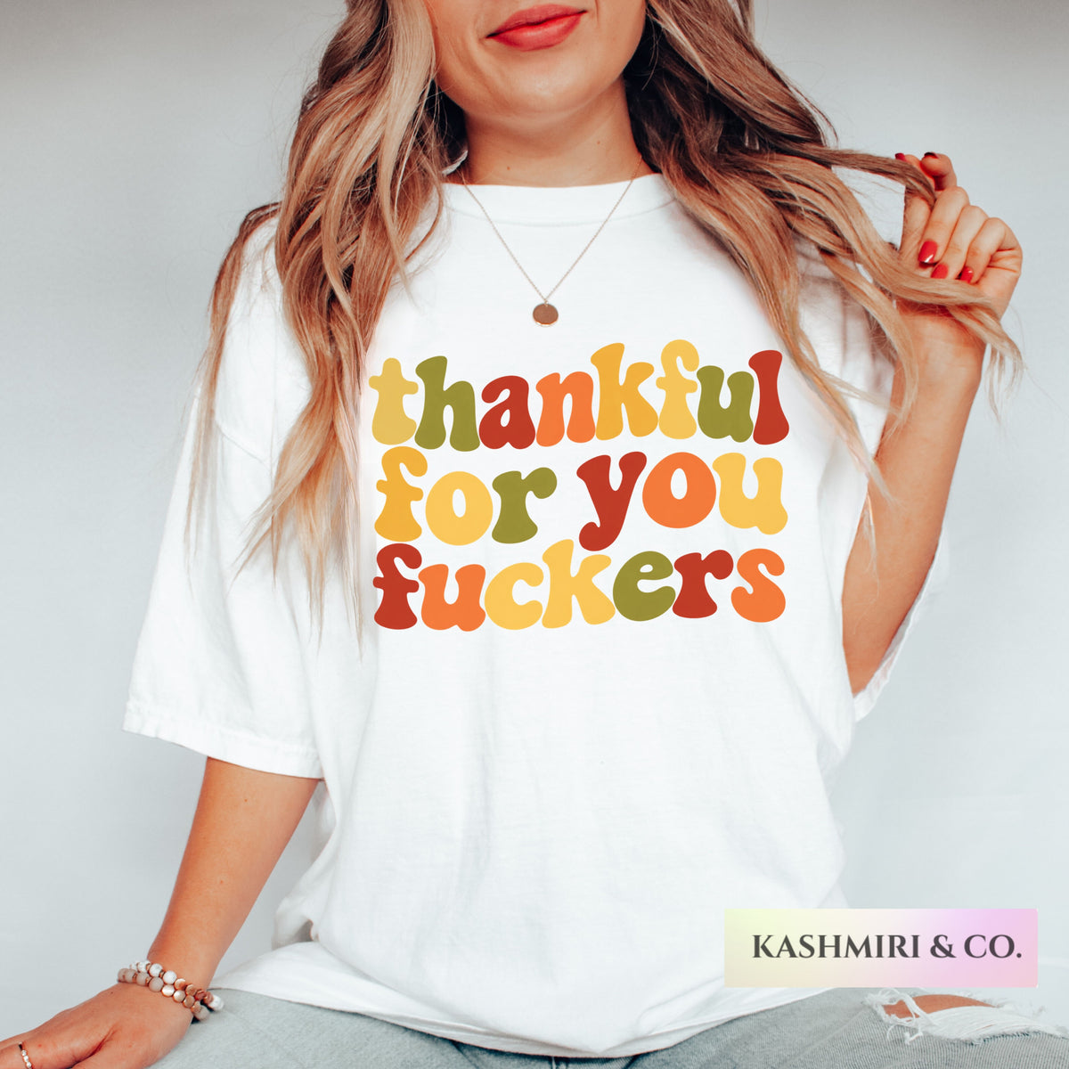 A woman wearing a white graphic t-shirt with the text 'thankful for you fuckers' printed in a retro, wavy multicolor font. The photo is cropped from the chin down, showing her hands, hair, and jeans against a plain light gray background.