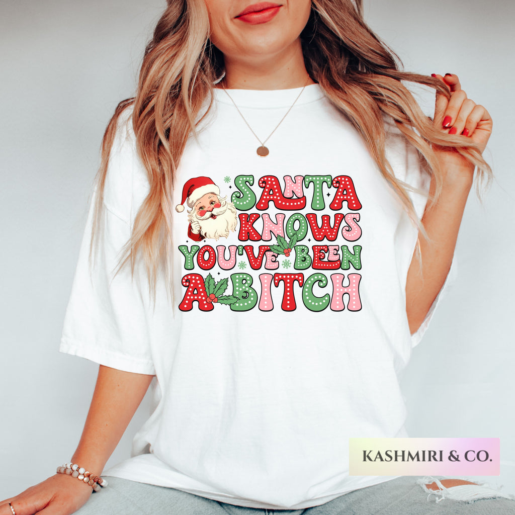 A woman wearing a white graphic t-shirt with a festive Christmas design that reads 'SANTA KNOWS YOU'VE BEEN A BITCH' in red and green glitter-style lettering with a Santa Claus illustration. The brand name 'KASHMIRI & CO.