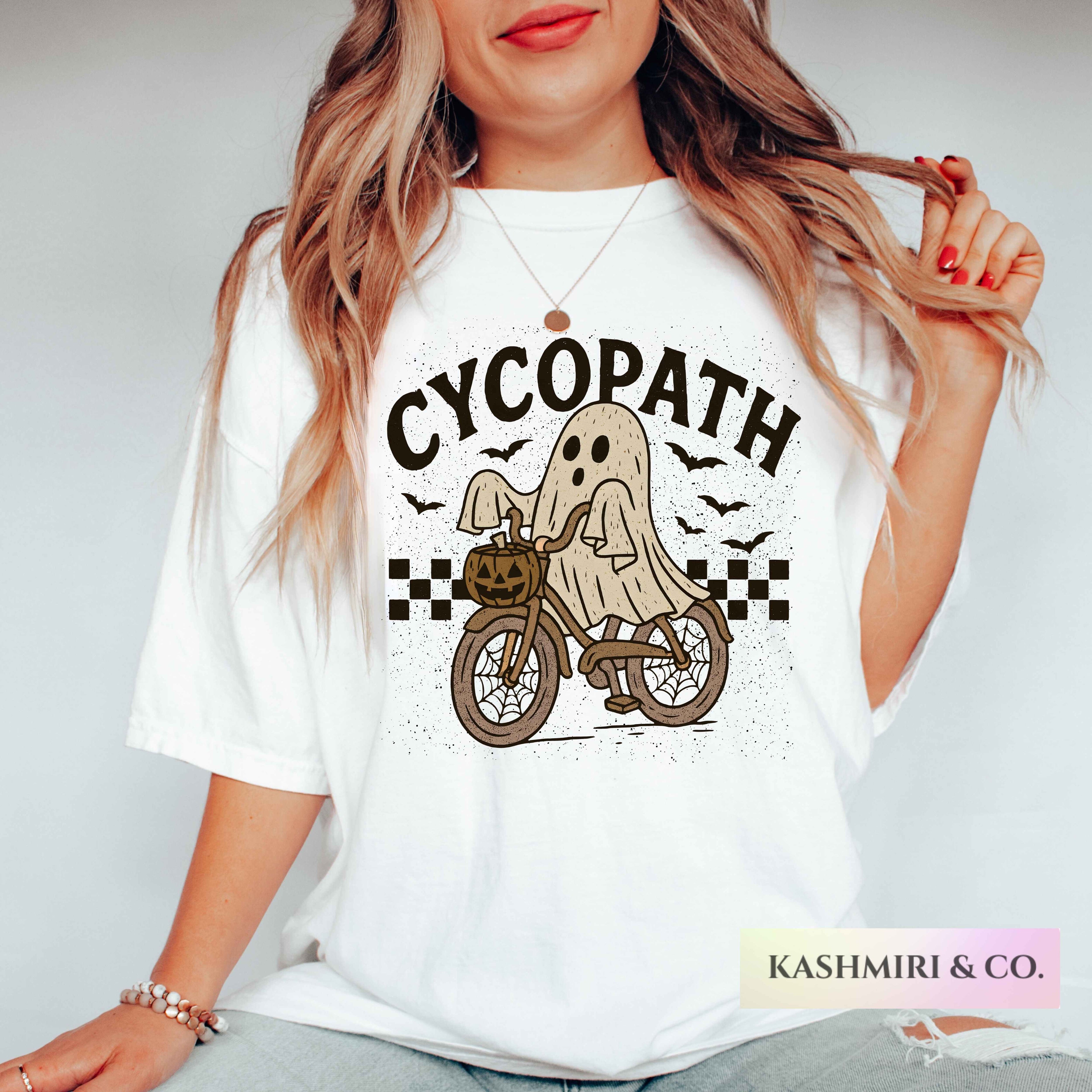 A woman wearing a white graphic t-shirt with a 'Cycopath' Halloween design featuring a ghost on a bicycle. The brand name 'Kashmiri & Co.