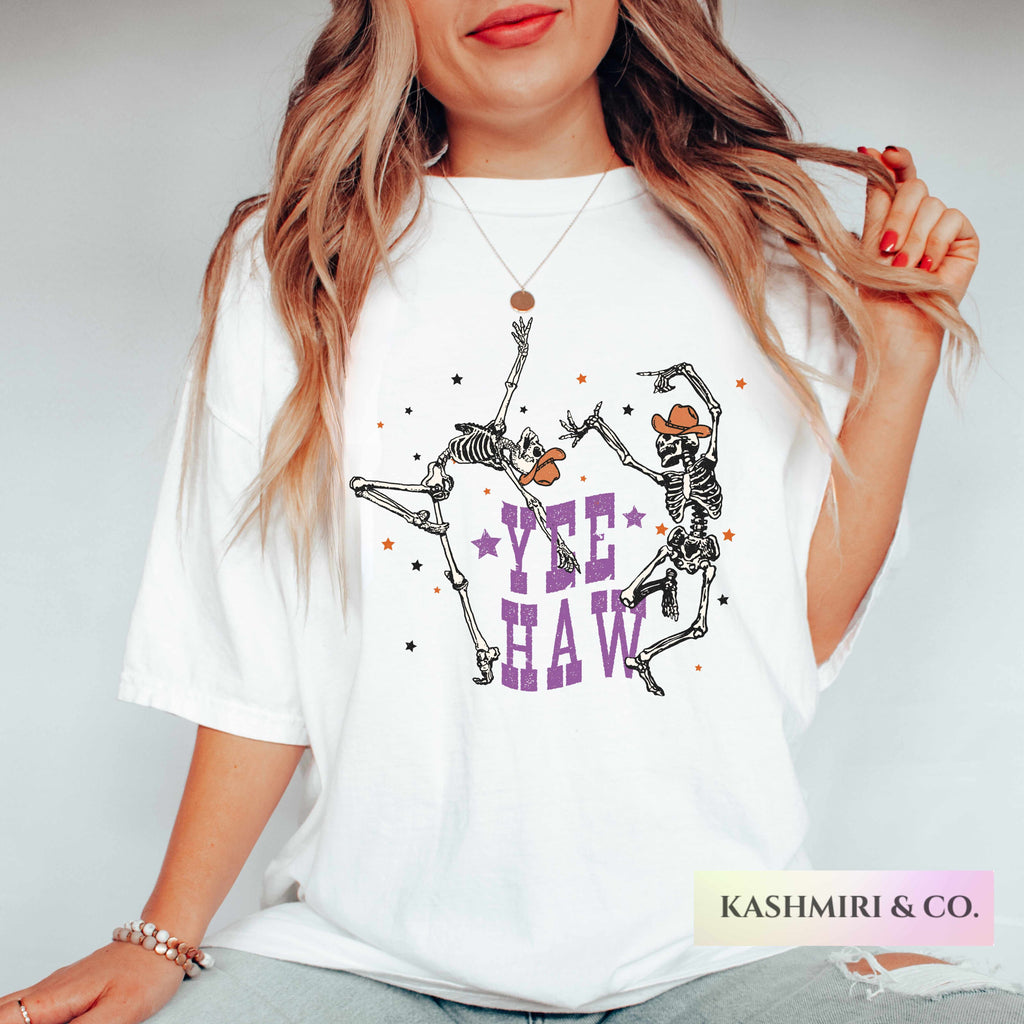A woman wearing a white oversized graphic T-shirt with two dancing skeleton figures and the text 'YEE HAW' in purple, shown against a light gray studio background. A 'KASHMIRI & CO.