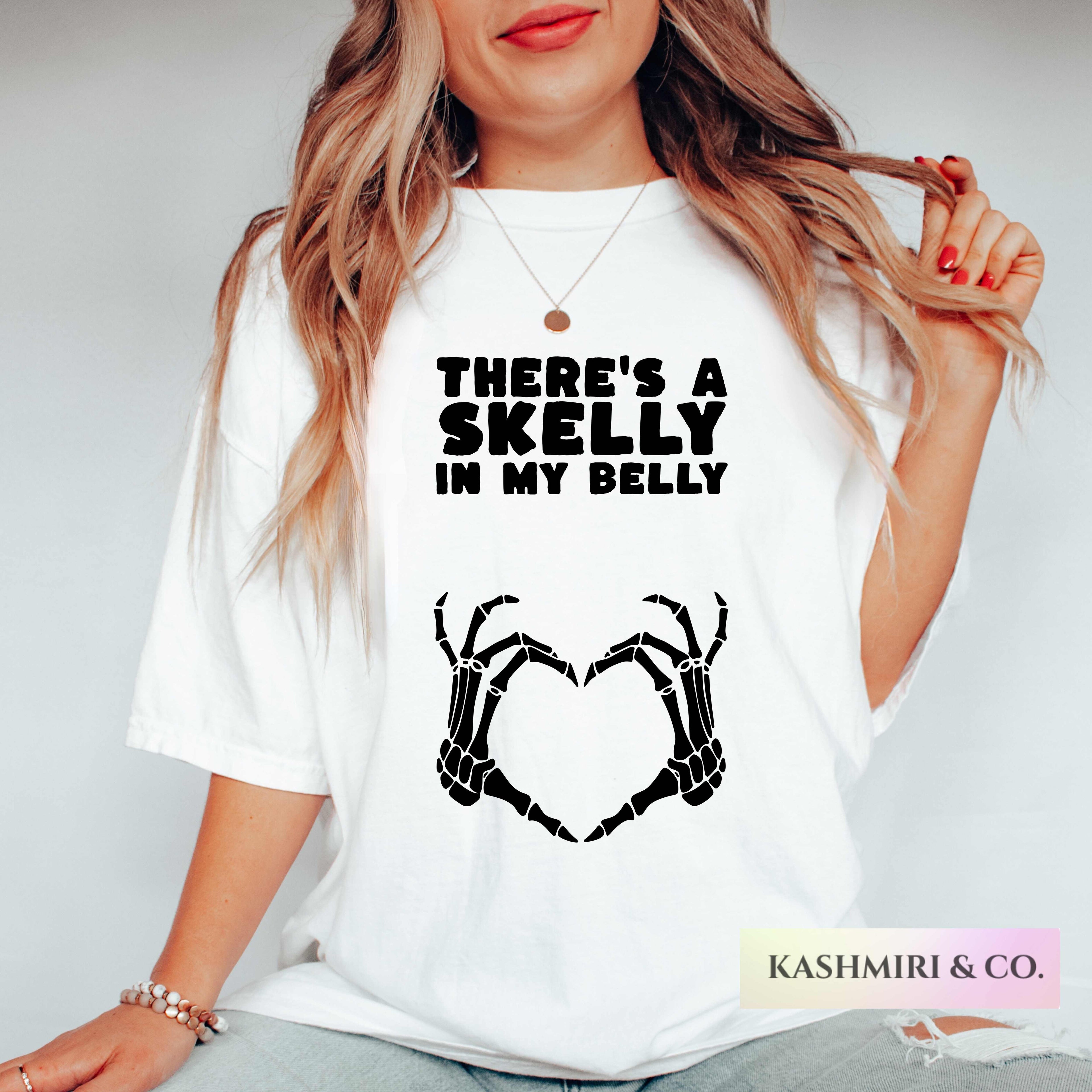 A woman wearing a white graphic t-shirt with the text 'THERE'S A SKELLY IN MY BELLY' and a black skeleton hand graphic forming a heart. The photo is cropped from the lips down, set against a plain light gray background.