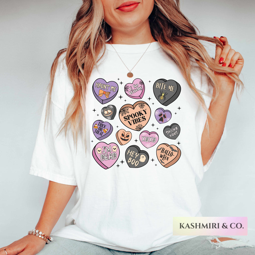 A woman wearing a white graphic t-shirt featuring a collection of colorful, spooky-themed heart designs with phrases like 'Cruel Love', 'Bite Me', and 'Spooky Vibes'. The brand name 'KASHMIRI & CO.