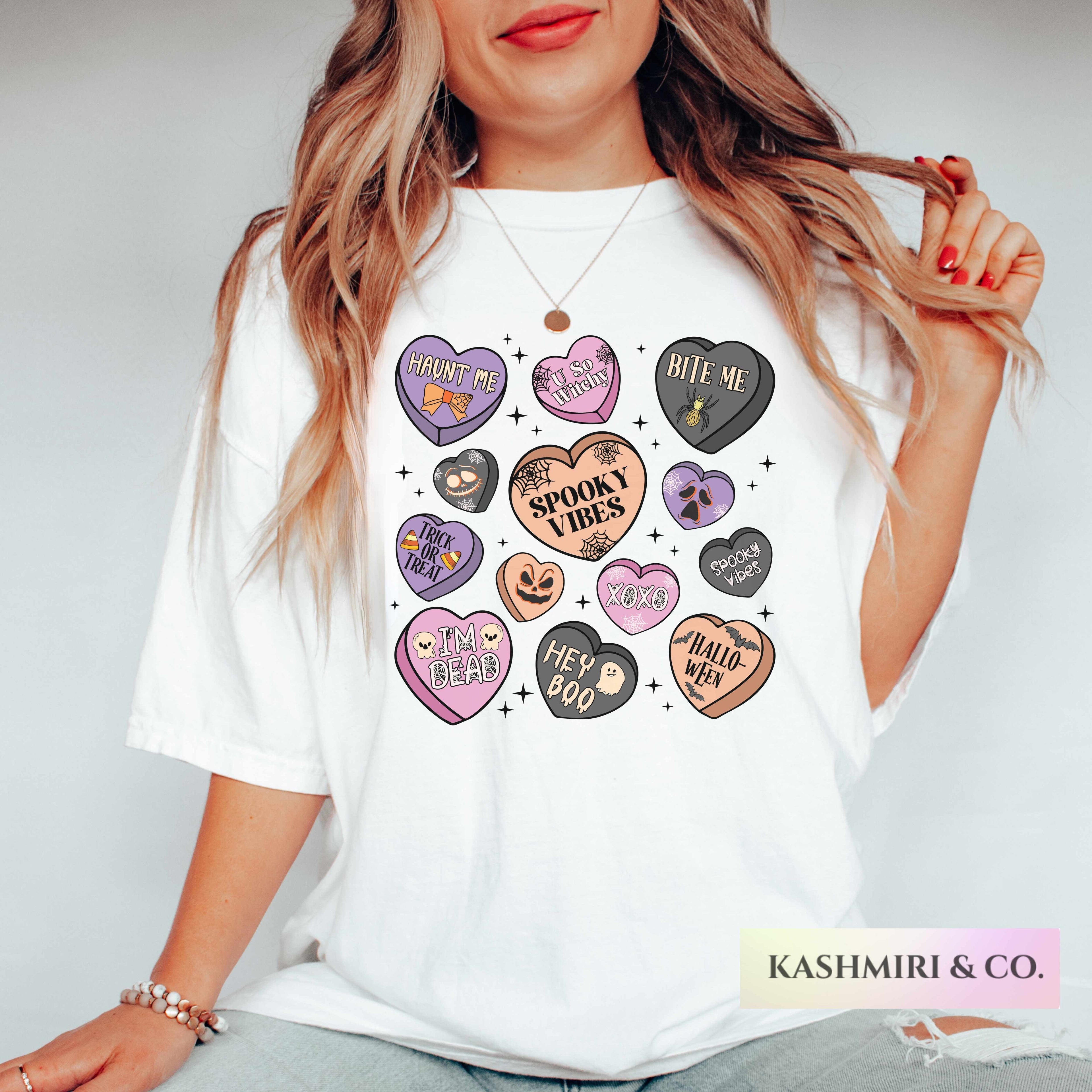 A woman wearing a white graphic t-shirt featuring a collection of colorful, spooky-themed heart designs with phrases like 'Cruel Love', 'Bite Me', and 'Spooky Vibes'. The brand name 'KASHMIRI & CO.