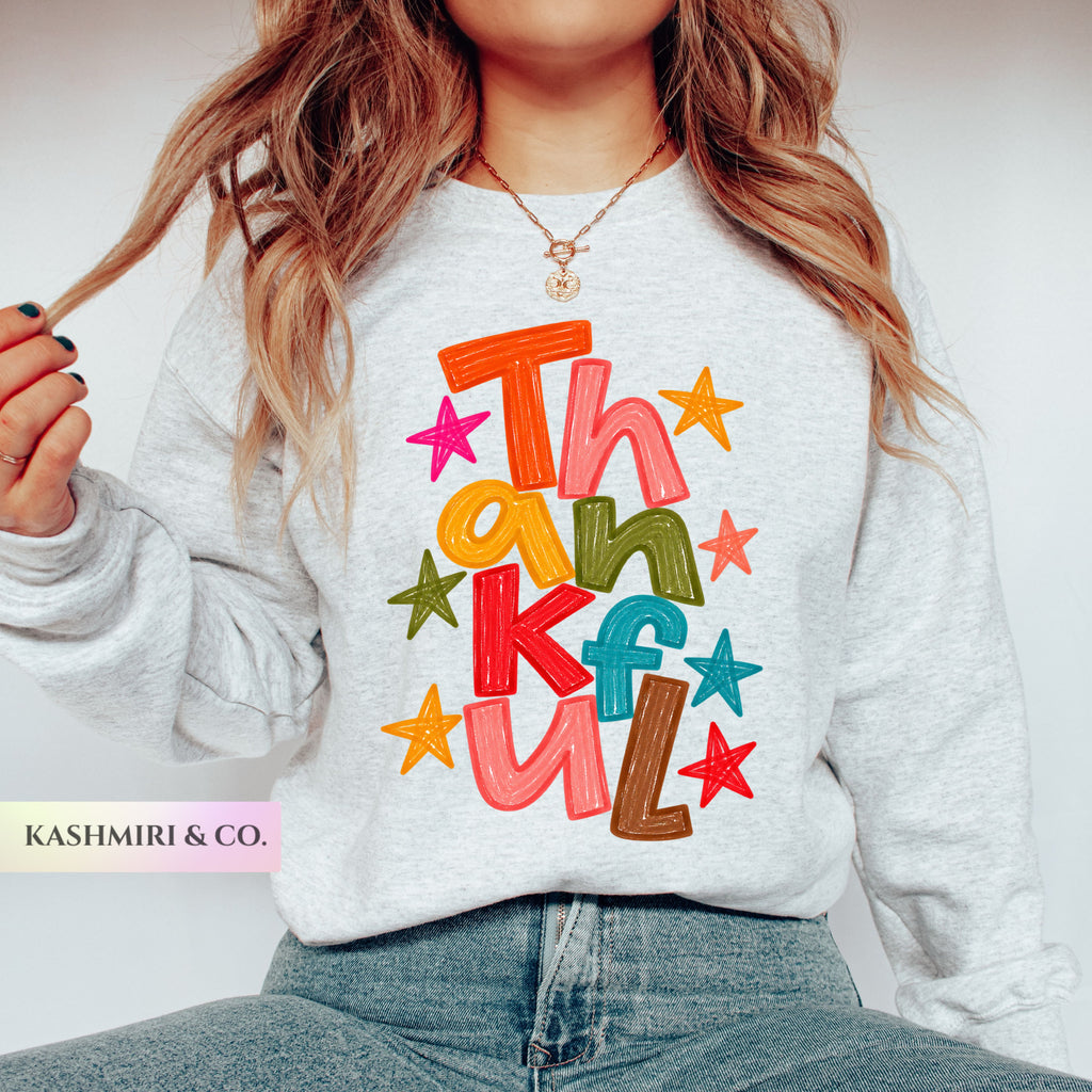 A woman wearing a light heather gray sweatshirt with a colorful 'Thankful' text and star graphic on the front, paired with blue jeans. The brand name 'KASHMIRI & CO.