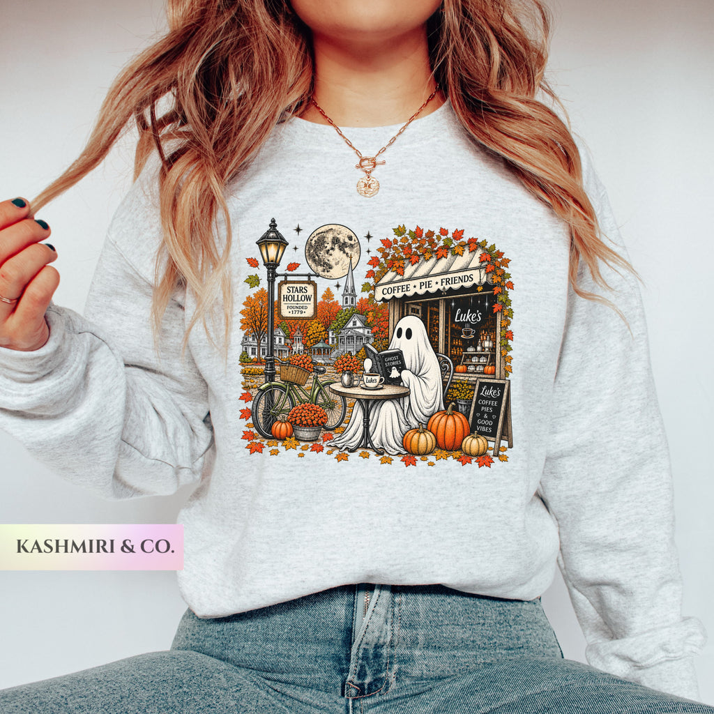 A woman wearing a light heather gray sweatshirt with a detailed autumn-themed graphic print featuring a ghost, pumpkins, a street lamp, and a moon. Stars Hallow Luke's Diner