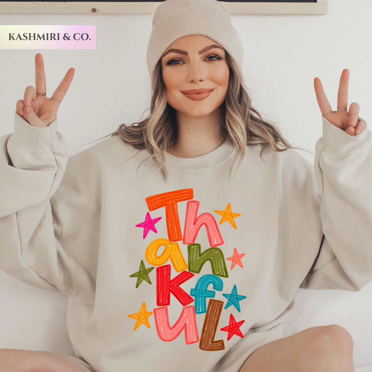 A woman wearing a beige knit beanie and a matching beige sweatshirt with a colorful 'Thankful' text and star graphic on the front, posing with peace signs against a light background. The brand name 'KASHMIRI & CO.