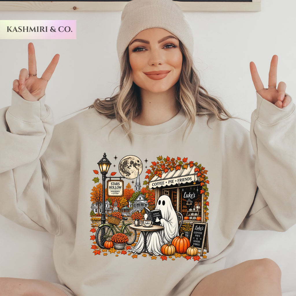 A woman wearing a light heather gray sweatshirt with a detailed autumn-themed graphic print featuring a ghost, pumpkins, a street lamp, and a moon. Stars Hallow Luke's Diner