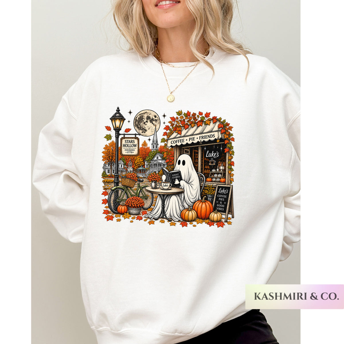 A woman wearing a light heather gray sweatshirt with a detailed autumn-themed graphic print featuring a ghost, pumpkins, a street lamp, and a moon. Stars Hallow Luke's Diner