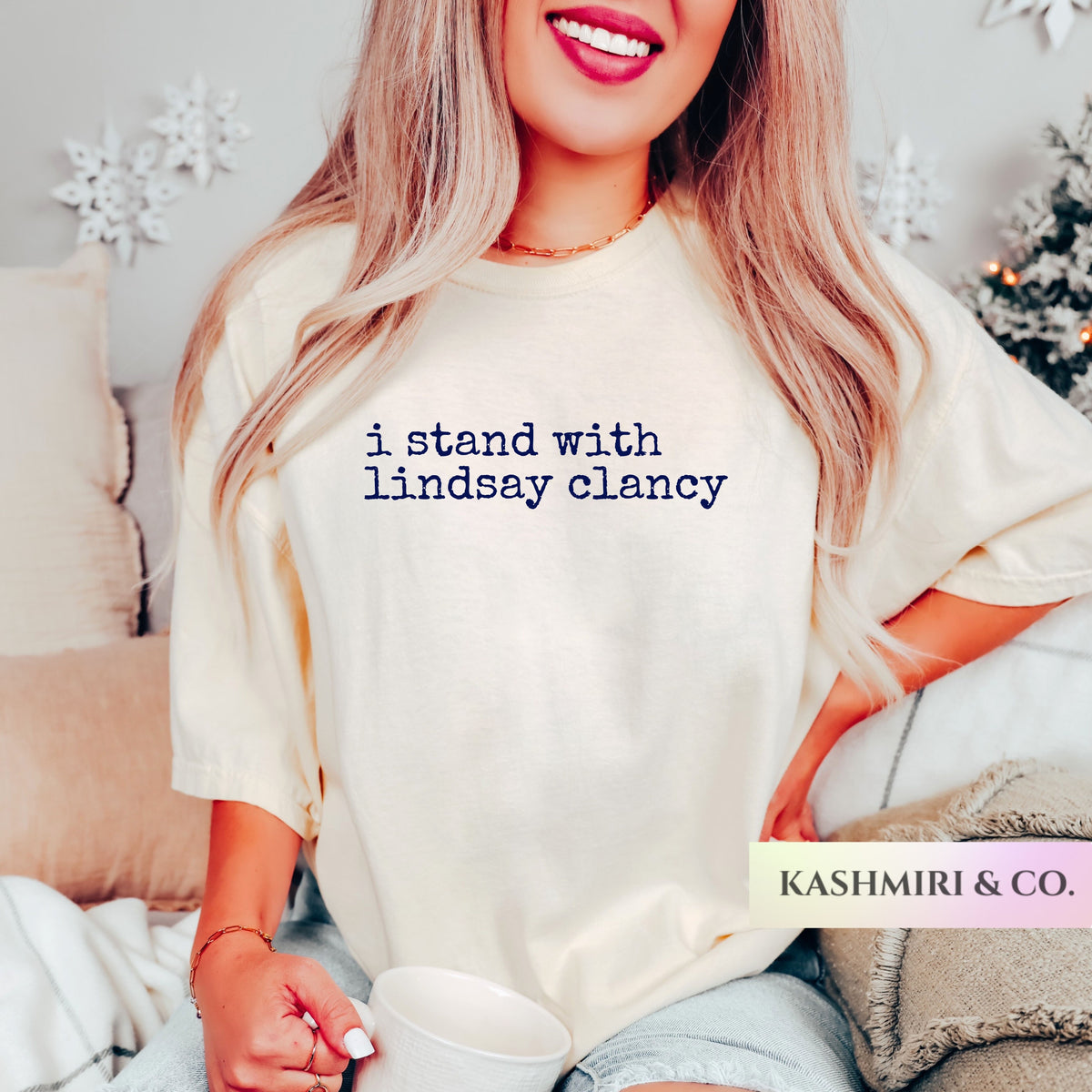 A woman wearing a cream-colored oversized t-shirt with the text 'i stand with lindsay clancy' printed in navy blue. She is holding a white mug in a cozy indoor setting with holiday decorations in the background.