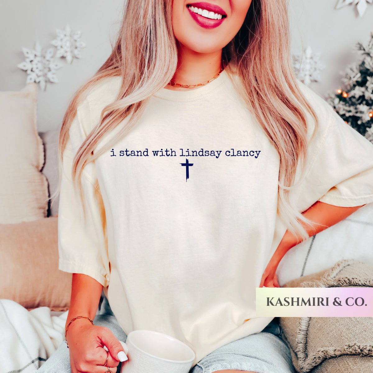 A woman wearing a cream-colored oversized t-shirt with the text 'i stand with lindsay clancy' and a cross printed in navy blue. She is holding a white mug in a cozy, decorated living room setting.