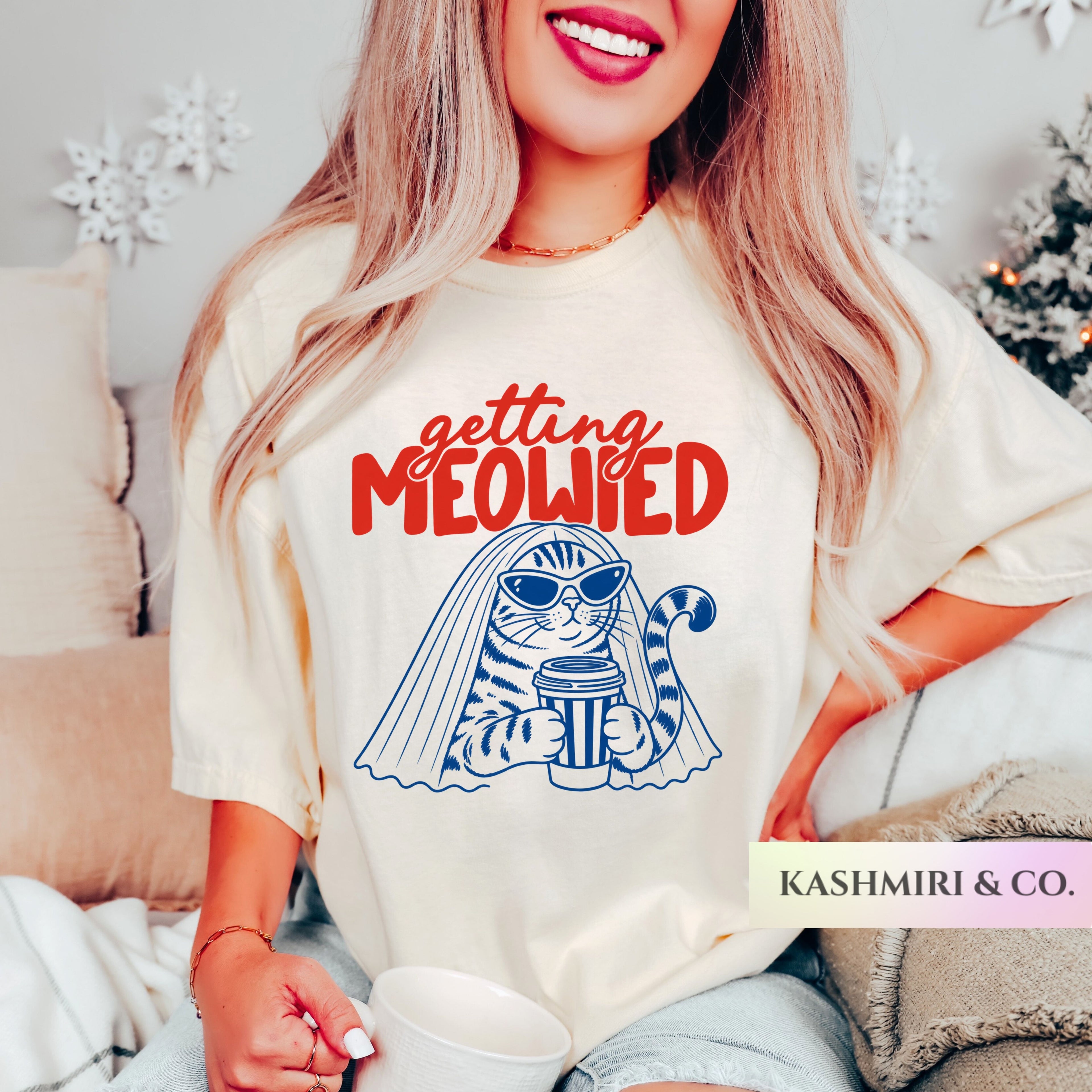 A woman wearing a cream-colored graphic t-shirt with a red and blue 'getting MEOWIED' cat illustration, sitting in a cozy indoor setting. The brand name 'KASHMIRI & CO.