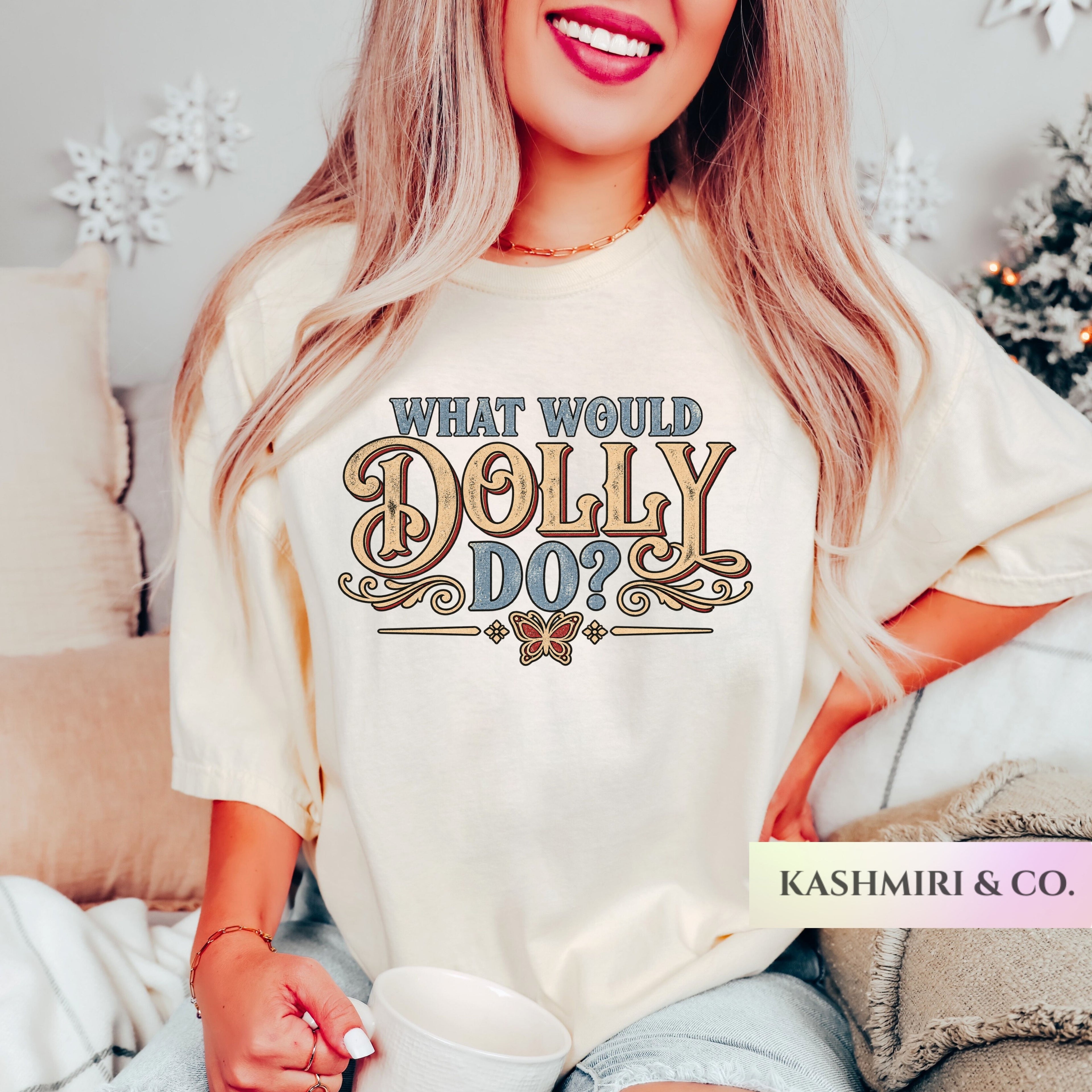 A woman wearing a cream-colored graphic t-shirt with the text 'WHAT WOULD DOLLY DO?' in stylized brown and blue lettering. She is holding a white mug in a cozy indoor setting with a bed and holiday decorations in the background.