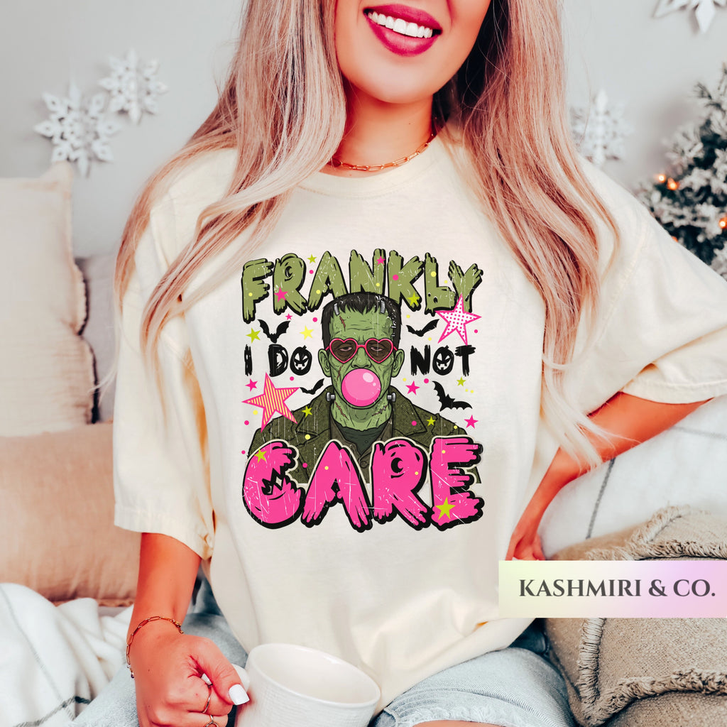A woman wearing a cream-colored graphic t-shirt with a Frankenstein-themed print that reads 'FRANKLY I DO NOT CARE' in green and pink lettering. The photo is taken in a cozy indoor setting with holiday decorations in the background, and a 'KASHMIRI & CO.