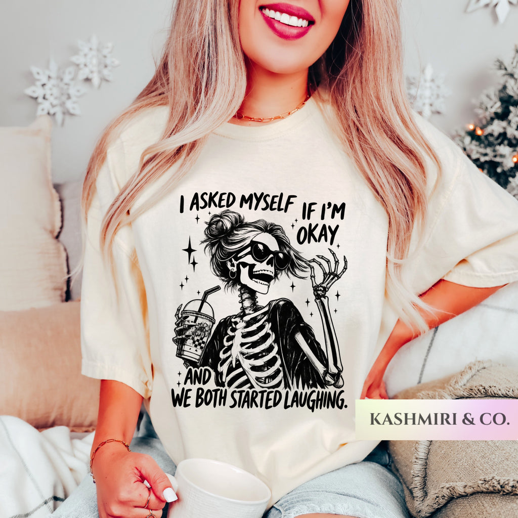 A woman wearing a cream-colored graphic t-shirt with a black skeleton drinking from a cup and the text 'I ASKED MYSELF IF I'M OKAY AND WE BOTH STARTED LAUGHING.' She is holding a white mug in a cozy indoor setting.