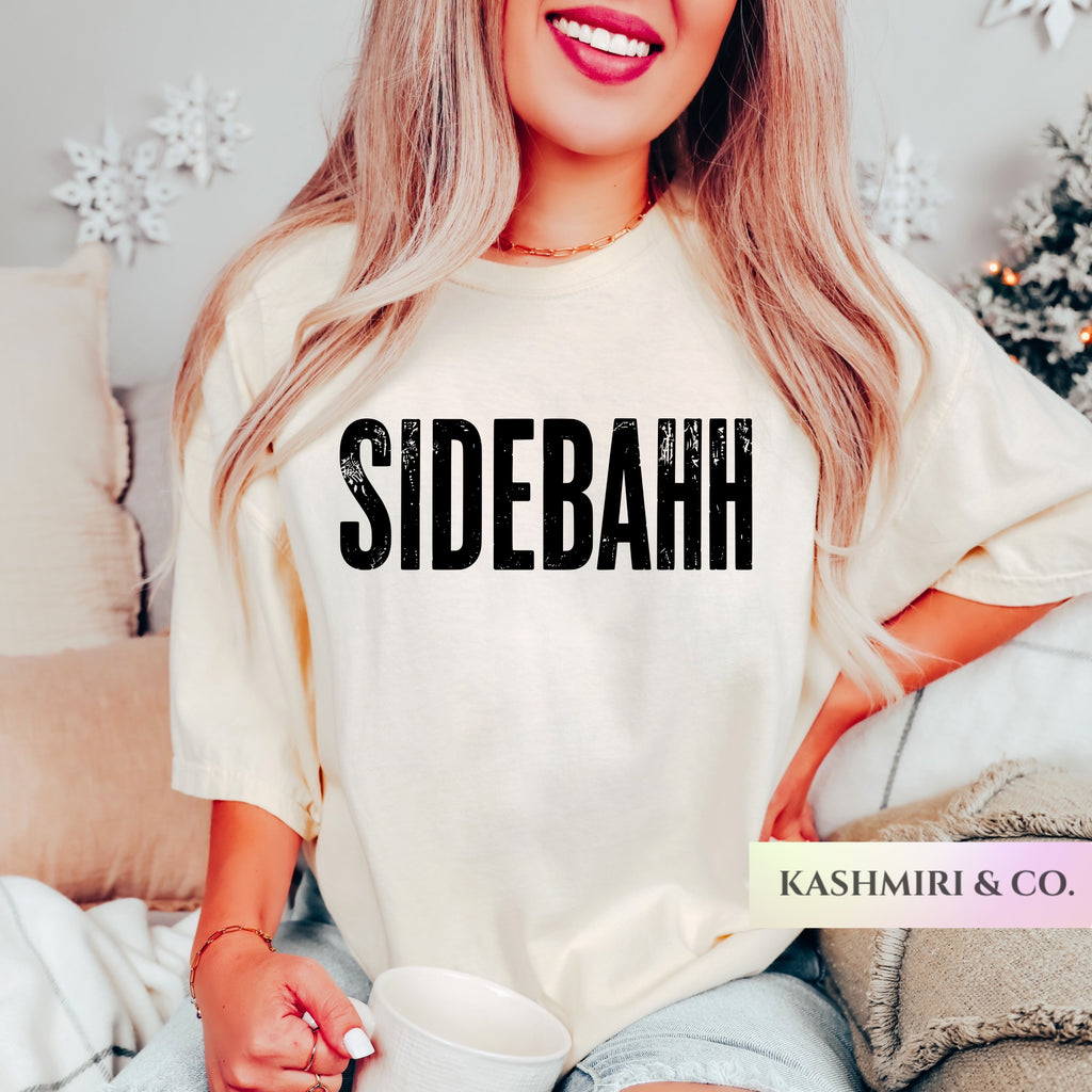 A woman wearing a cream-colored oversized t-shirt with the word 'SIDEBAHH' printed in bold black letters across the chest. She is holding a white mug in a cozy indoor setting with pillows and holiday decorations in the background.