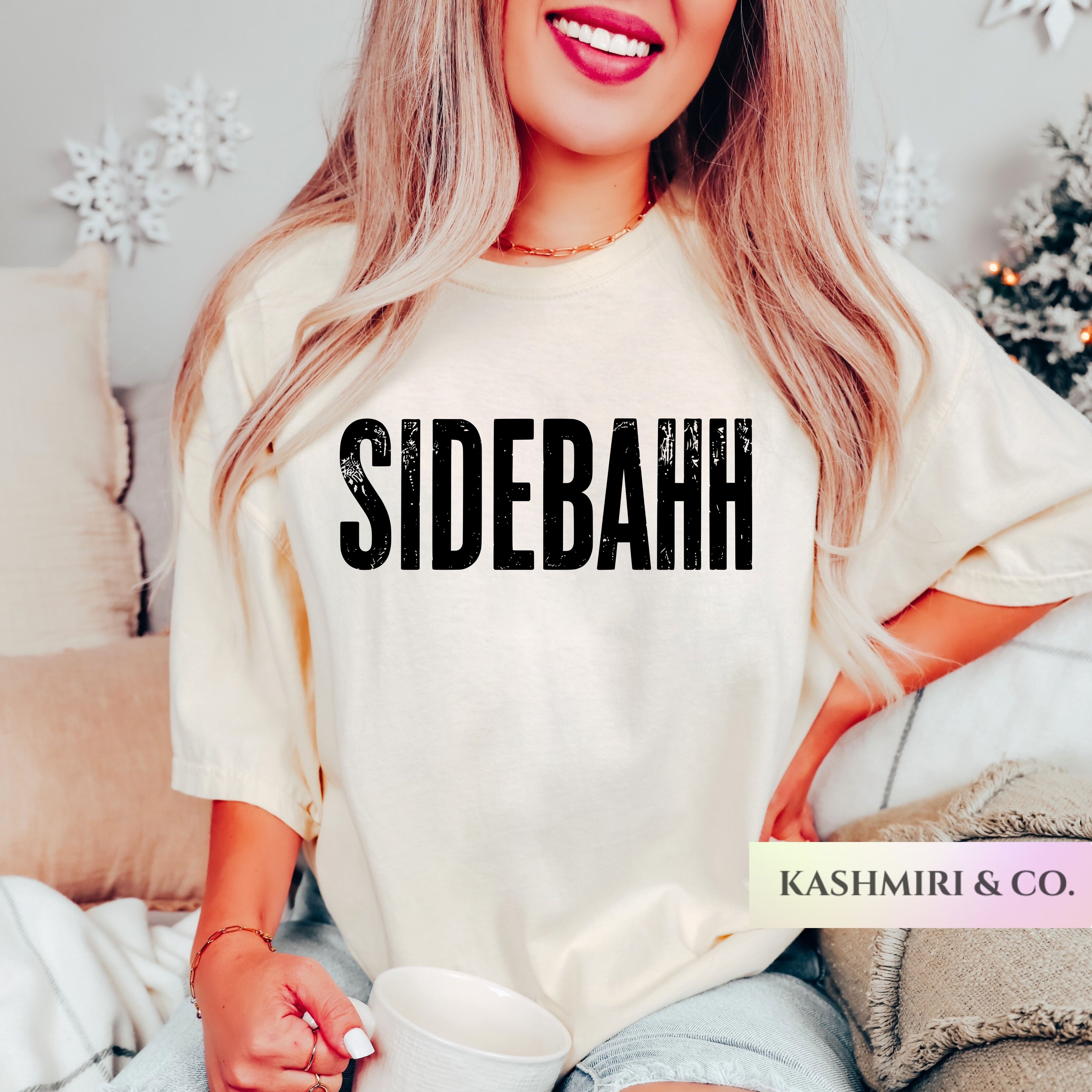 A woman wearing a cream-colored oversized t-shirt with the word 'SIDEBAHH' printed in bold black letters across the chest. She is holding a white mug in a cozy indoor setting with pillows and holiday decorations in the background.