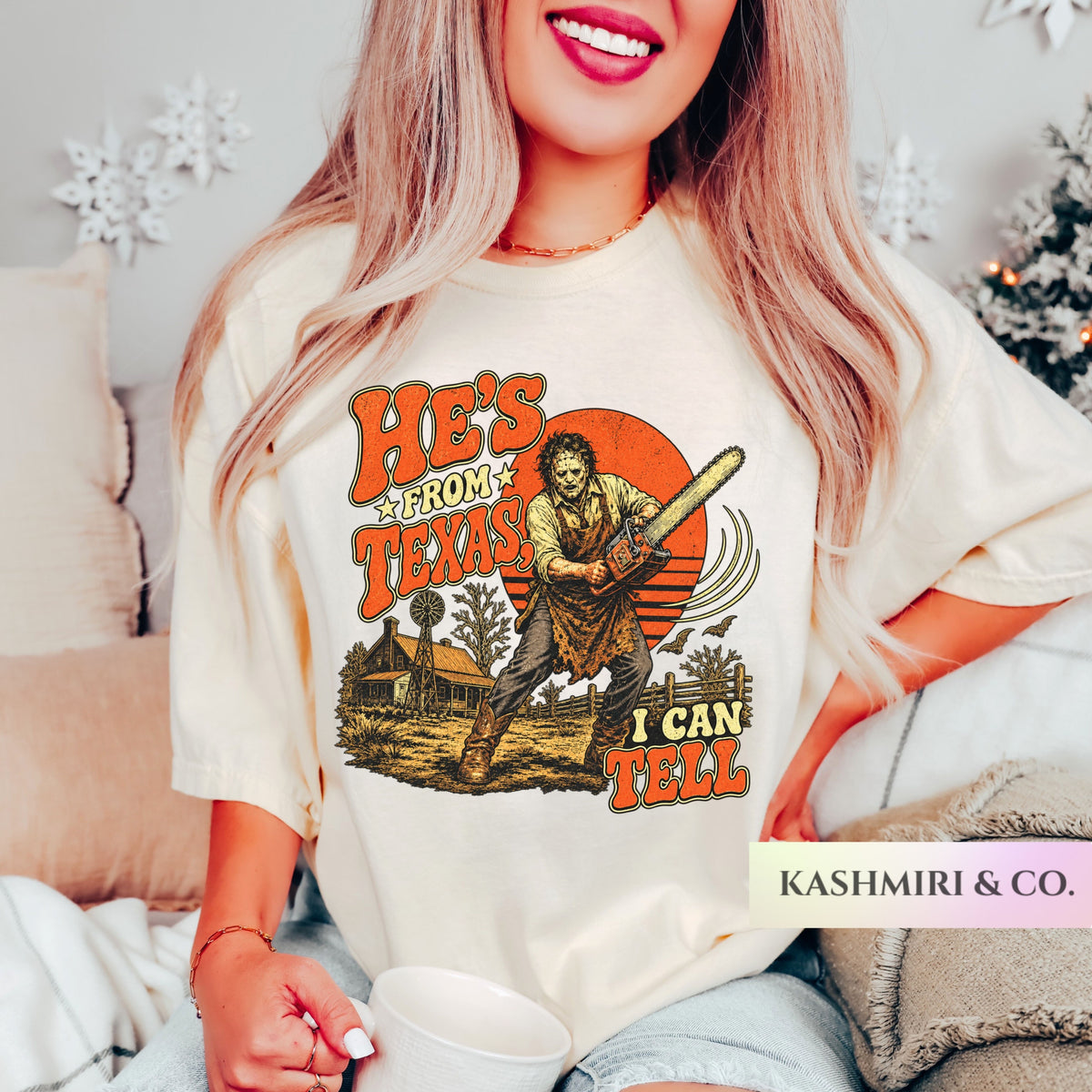 A woman wearing an oversized cream graphic t-shirt with a horror-themed print featuring Leatherface and the text 'HE'S FROM TEXAS I CAN TELL'. The photo is taken in a cozy bedroom setting with a bed, pillows, and a Christmas tree in the background.