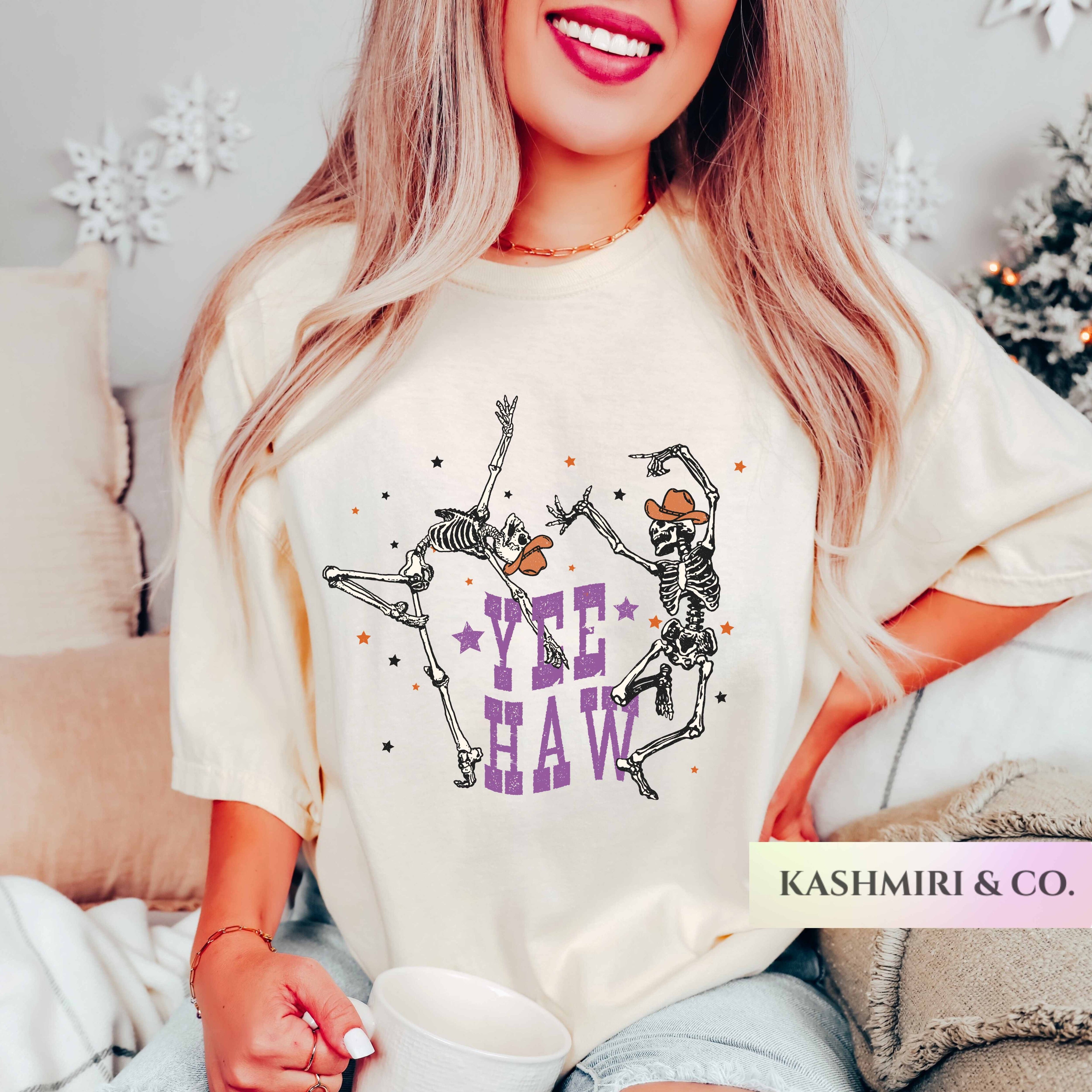 A woman wearing a cream-colored graphic t-shirt with a skeleton dancing design and the text 'YEE HAW' in purple. She is sitting in a cozy room setting, holding a white mug.