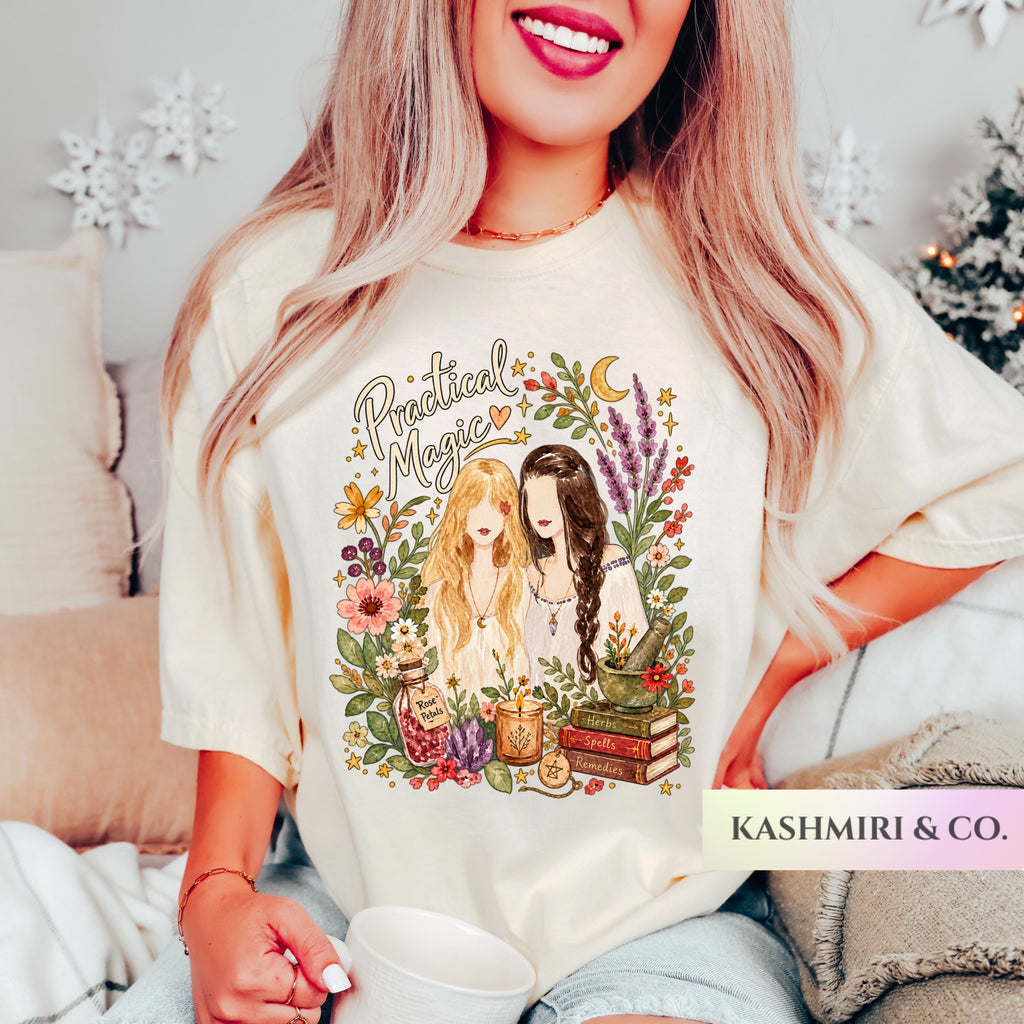 A woman wearing a cream-colored graphic t-shirt with a 'Practical Magic' illustration featuring two girls, books, and flowers. The t-shirt is shown in a cozy bedroom setting with a blanket and holiday decorations in the background.