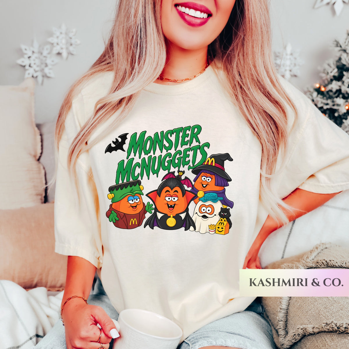 A woman wearing a cream-colored graphic t-shirt with a 'Monster McNuggets' cartoon design, sitting in a cozy indoor setting with a mug.