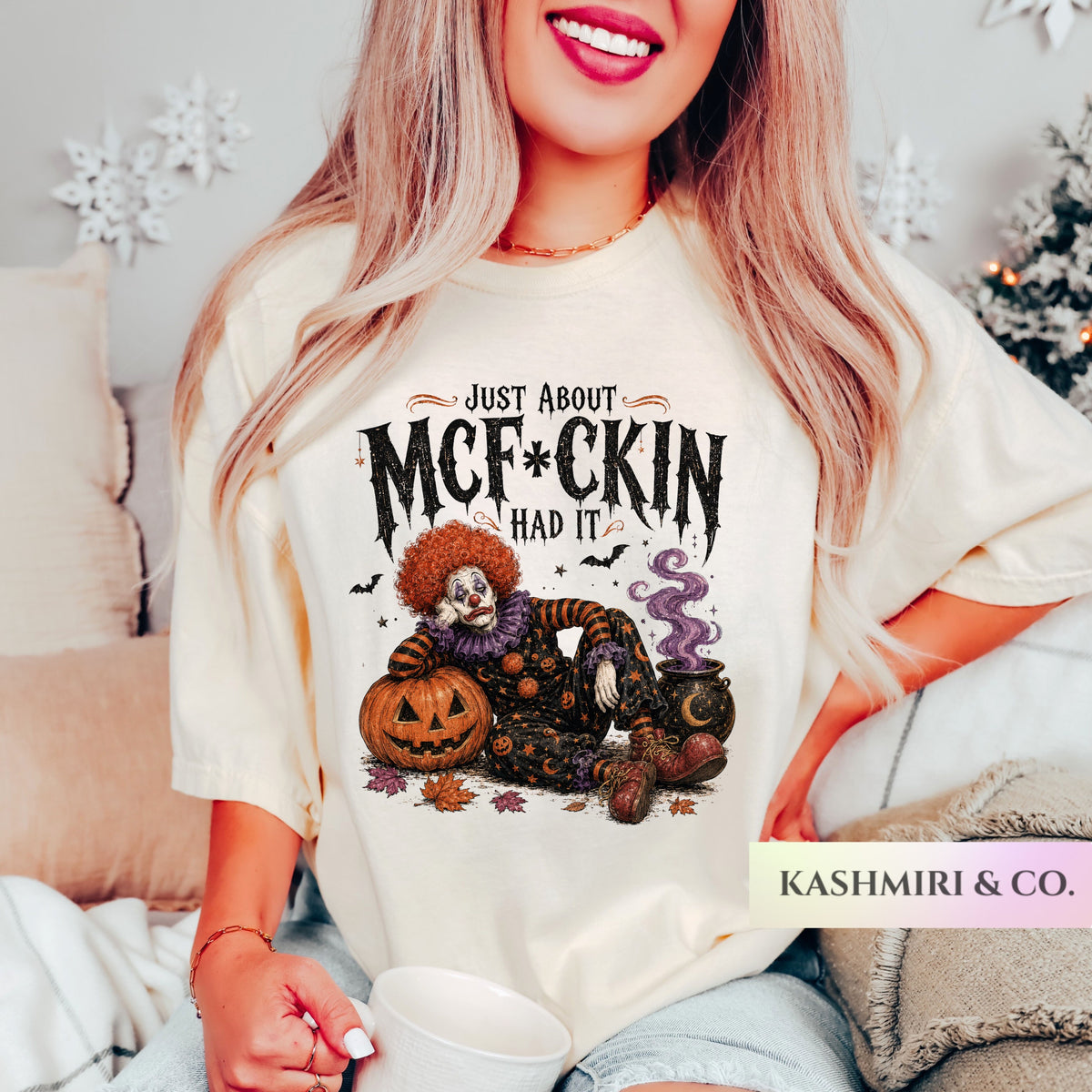 A woman wearing a cream-colored graphic t-shirt with a Halloween clown and jack-o'-lantern design and the text 'JUST ABOUT MCF*CKIN' HAD IT'. The photo is taken in a cozy bedroom setting with a bed, pillows, and holiday decorations in the background.