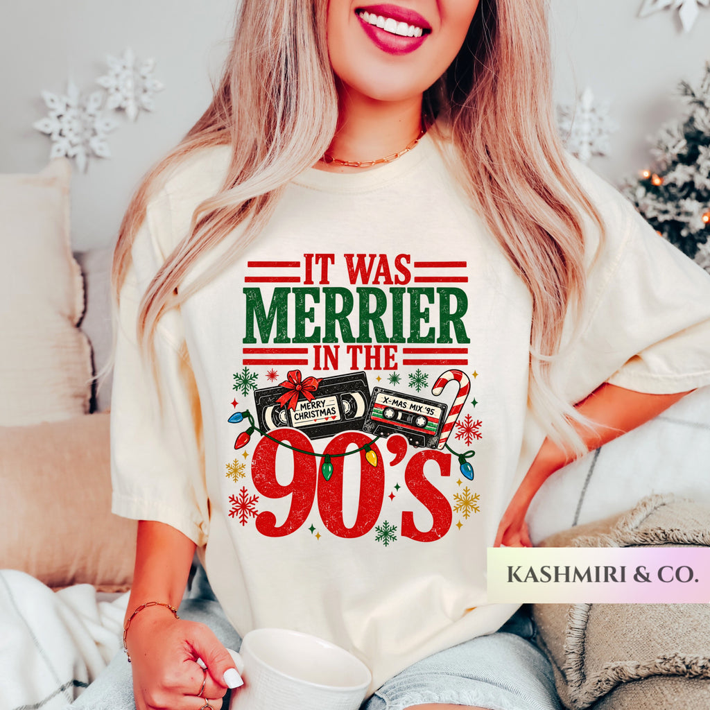 A woman wearing a cream-colored graphic t-shirt with a festive Christmas design that reads 'IT WAS MERRIER IN THE 90's' in red and green text, featuring cassette tapes, candy canes, and snowflakes. She is sitting in a cozy bedroom setting with a mug, and a 'KASHMIRI & CO.