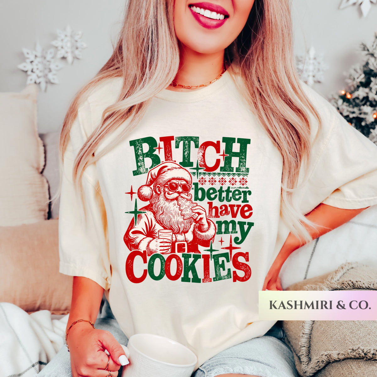 A woman wearing a cream-colored graphic t-shirt with a red and green Christmas-themed design that reads 'BITCH better have my COOKIES' alongside an illustration of Santa Claus. She is holding a white mug in a cozy indoor setting with holiday decorations in the background.