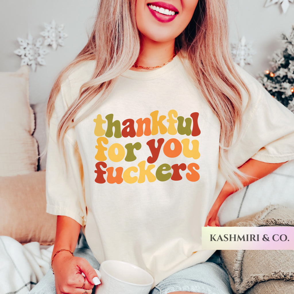 A woman wearing a cream-colored graphic t-shirt with the text 'thankful for you fuckers' printed in a retro, wavy multicolor font. She is holding a white mug in a cozy indoor setting with a Christmas tree in the background.