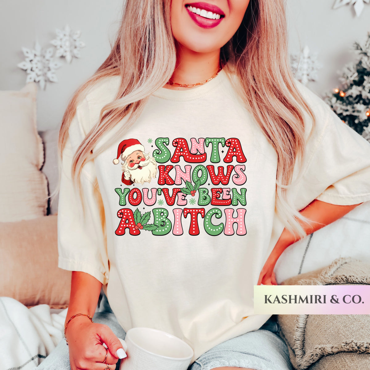 A woman wearing a cream-colored graphic t-shirt with a festive Christmas design that reads 'SALTA KNOWS YOU'VE BEEN A BITCH' in red and green glitter-style lettering with a Santa Claus illustration. The brand name 'KASHMIRI & CO.