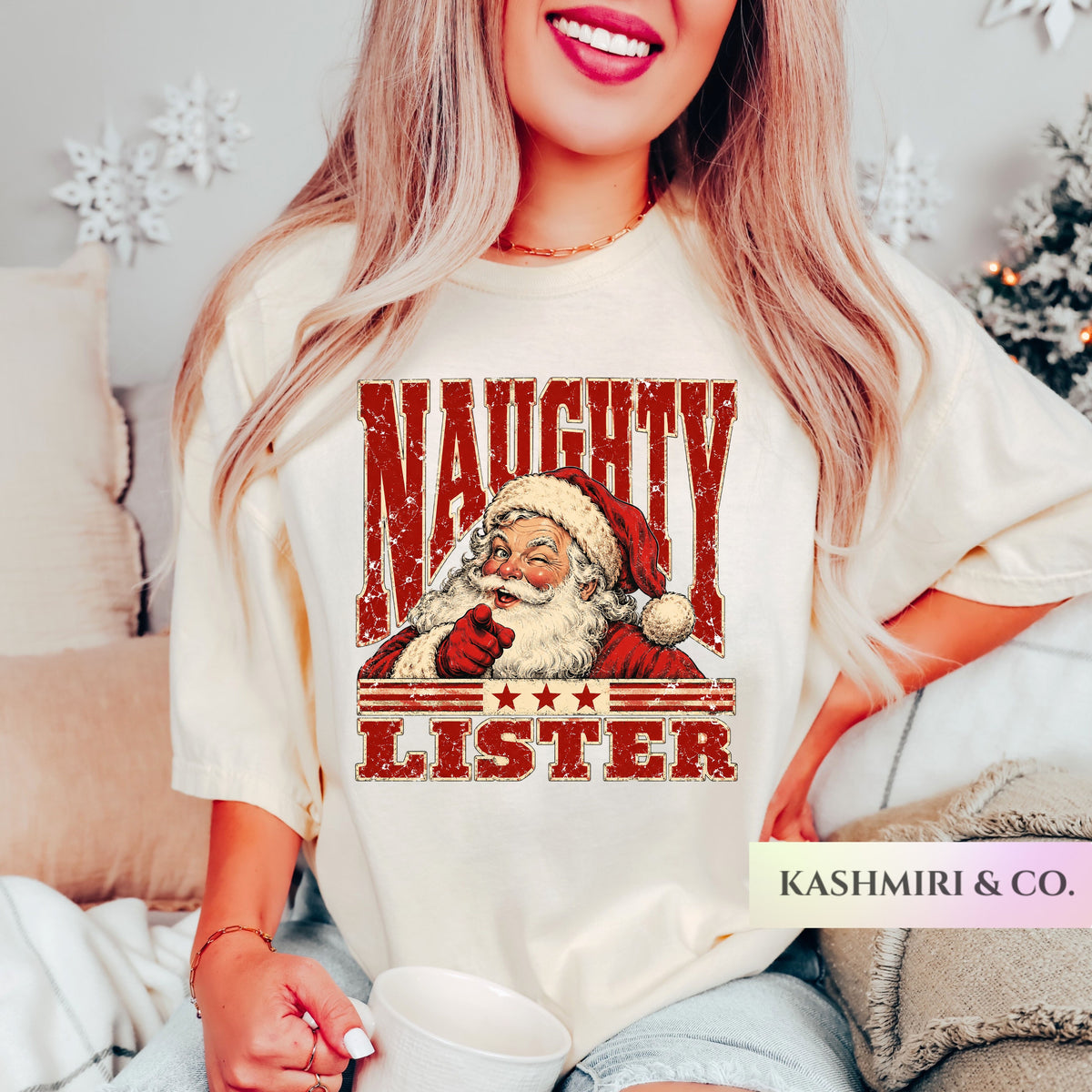 A woman wearing a cream-colored graphic t-shirt with a distressed red and brown print of Santa Claus and the text 'NAUGHTY LISTER'. The photo is taken in a cozy indoor setting with holiday decorations in the background, and a 'KASHMIRI & CO.