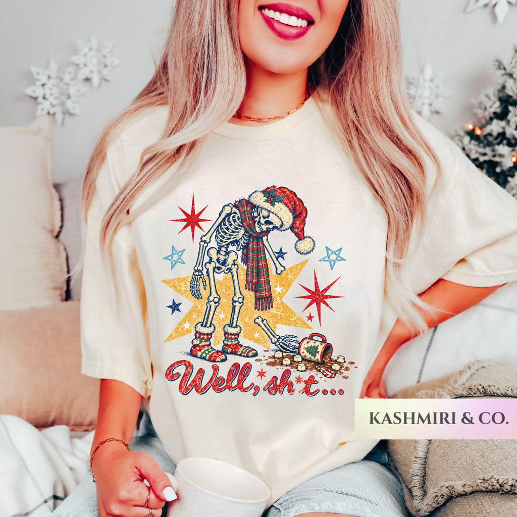 A woman wearing a cream-colored graphic t-shirt with a cartoon skeleton in a Santa hat and scarf, holding a white mug in a cozy indoor setting. The brand name 'KASHMIRI & CO.