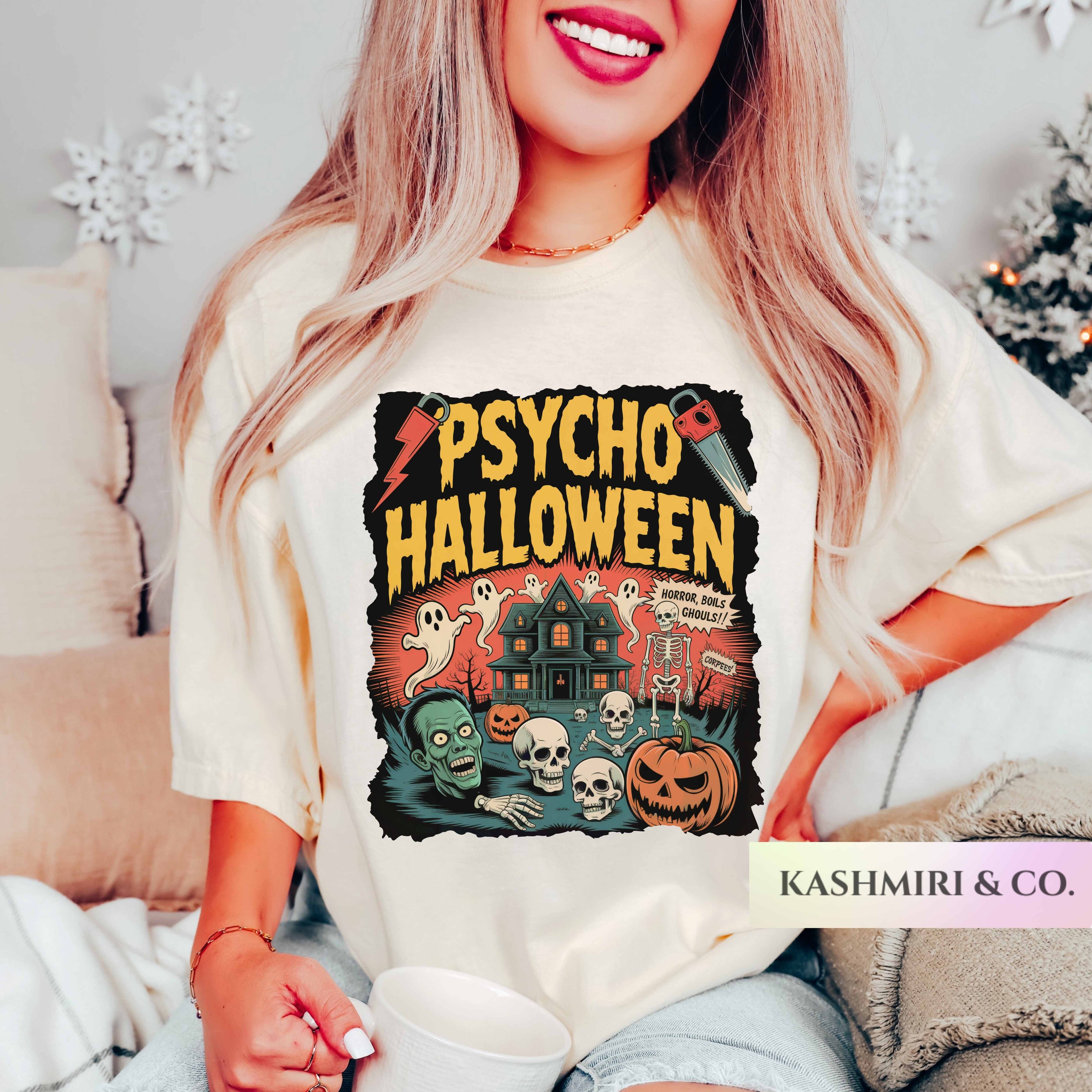A woman wearing a cream-colored graphic t-shirt with a 'Psycho Halloween' print, sitting in a cozy living room setting. The graphic features a haunted house, skulls, a pumpkin, and ghosts.