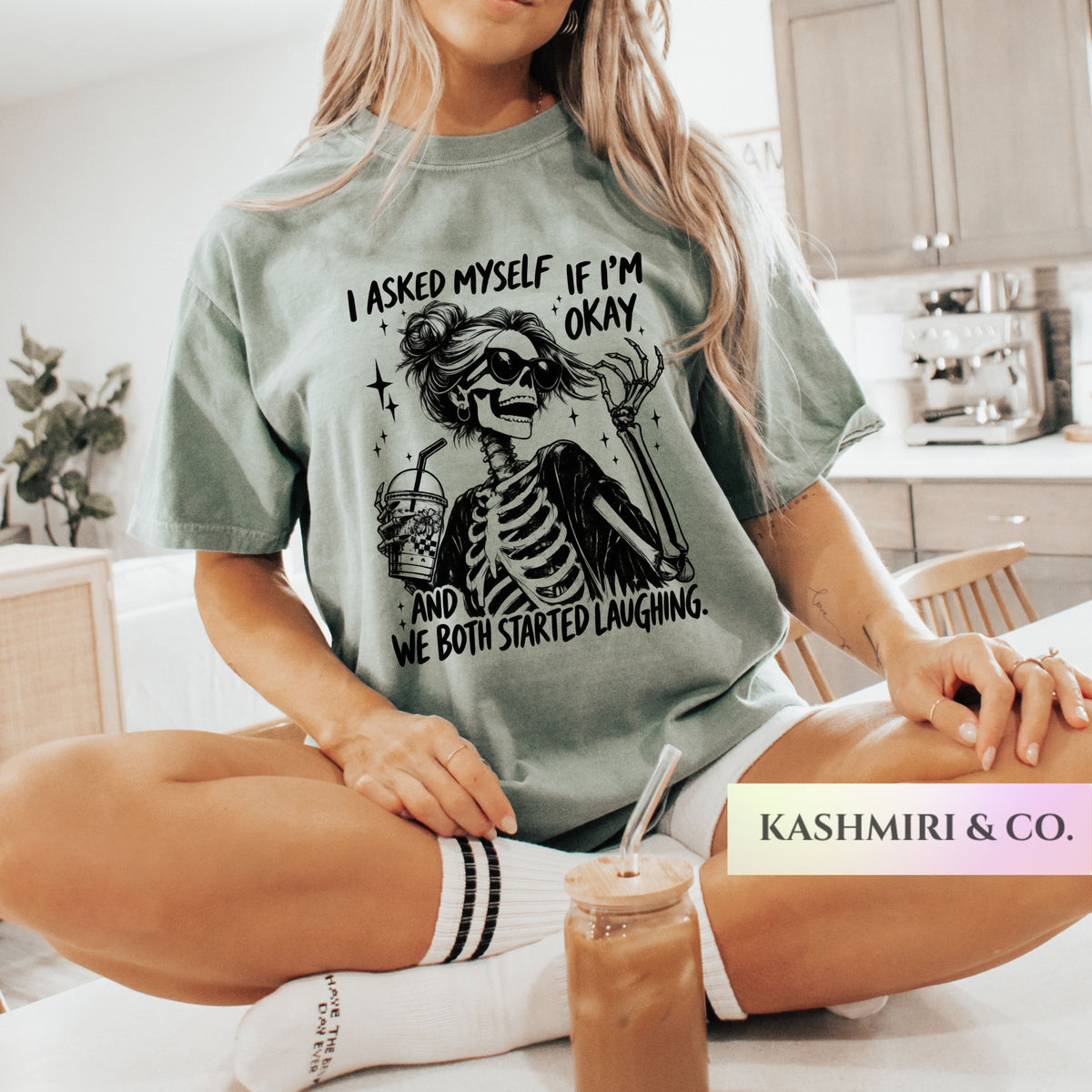 A woman sitting cross-legged in a kitchen setting wearing a sage green graphic t-shirt with a black skeleton meme design and text. A pink and white 'KASHMIRI & CO.